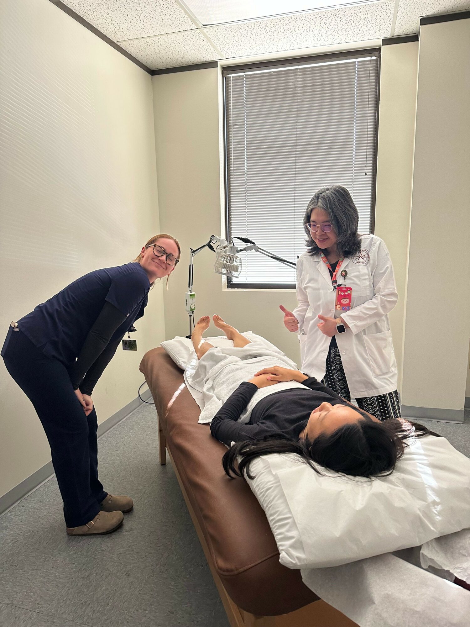 Person lying in hospital bed with a nurse and visitor nearby in a hospital room.