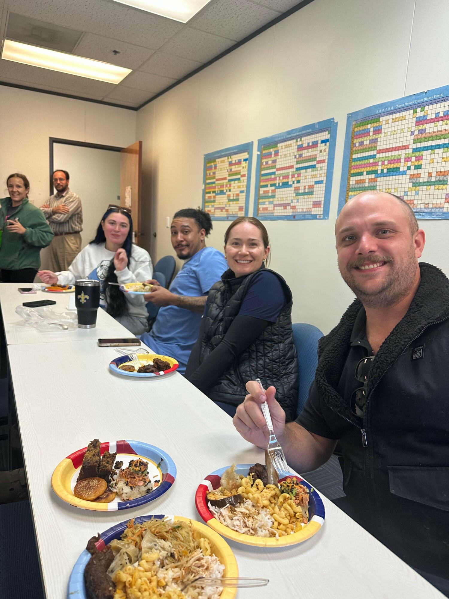 Four people sitting at a table with plates of food, smiling, in a room with colorful posters on the wall.
