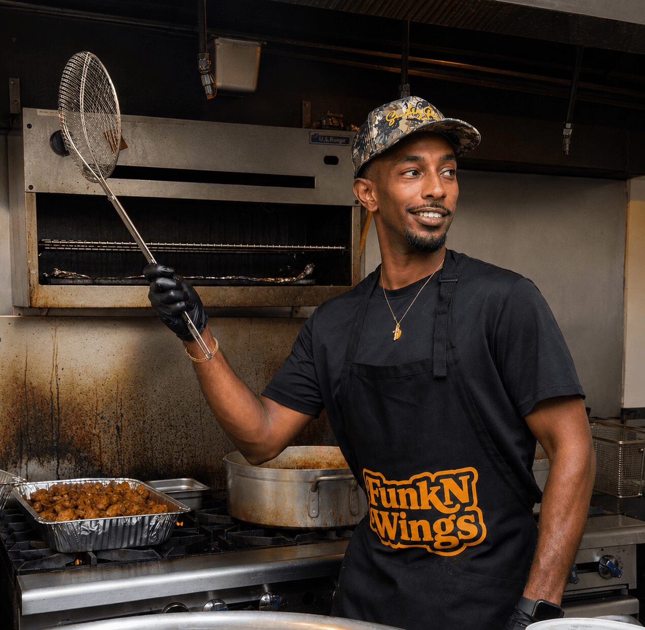 Man cooking in a kitchen, holding a frying net, wearing a black apron with 'FunkN Wings' logo, smiling, wearing a cap.