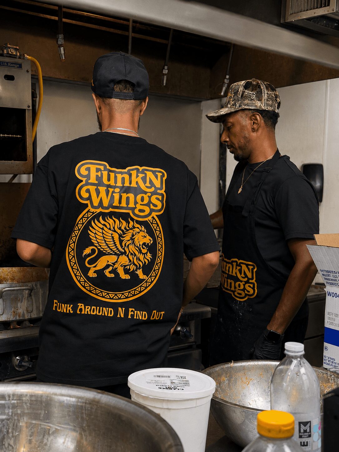 Two men in black shirts with FunkN Wings logo, one with a cap, in a kitchen setting with bowls and bottles.