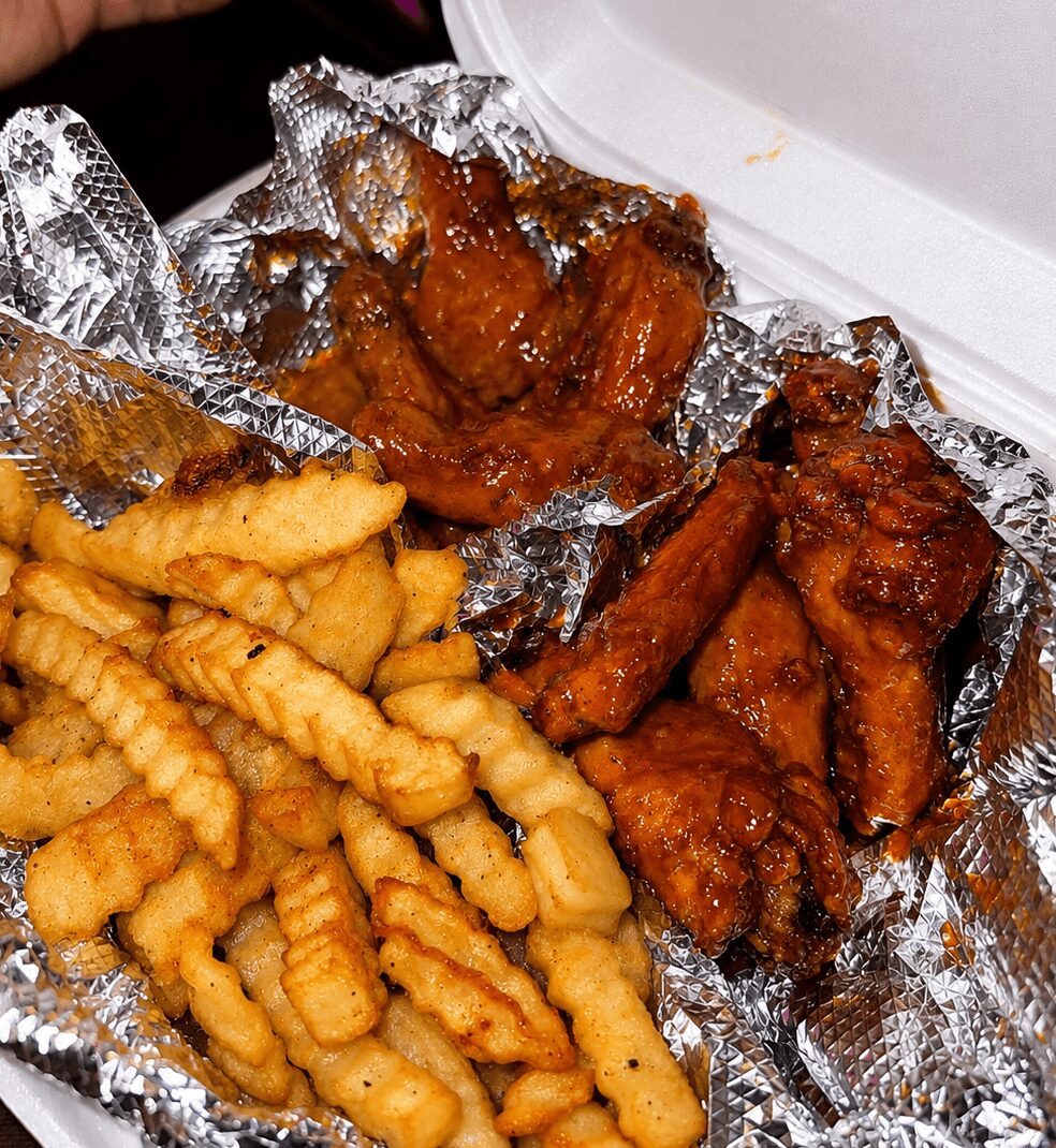 Fried crinkle-cut fries and glazed chicken wings in foil container, partially inside a white takeout box.