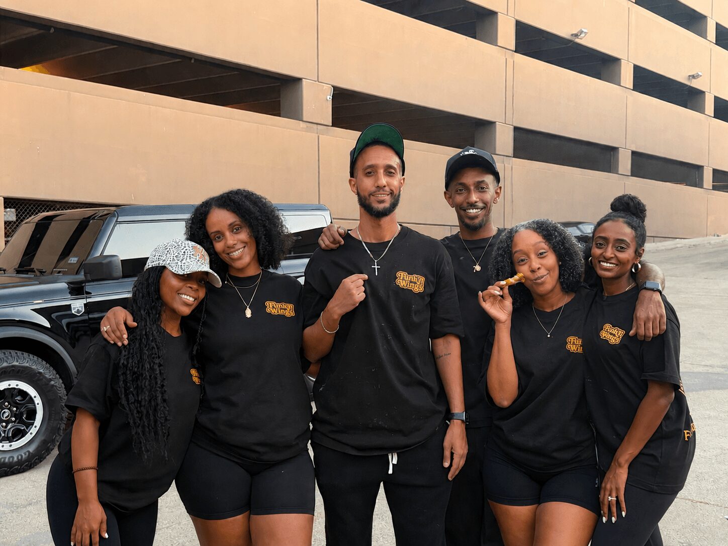Group of six people standing outdoors in front of a building, smiling, wearing matching black shirts, some holding food, with a vehicle nearby.