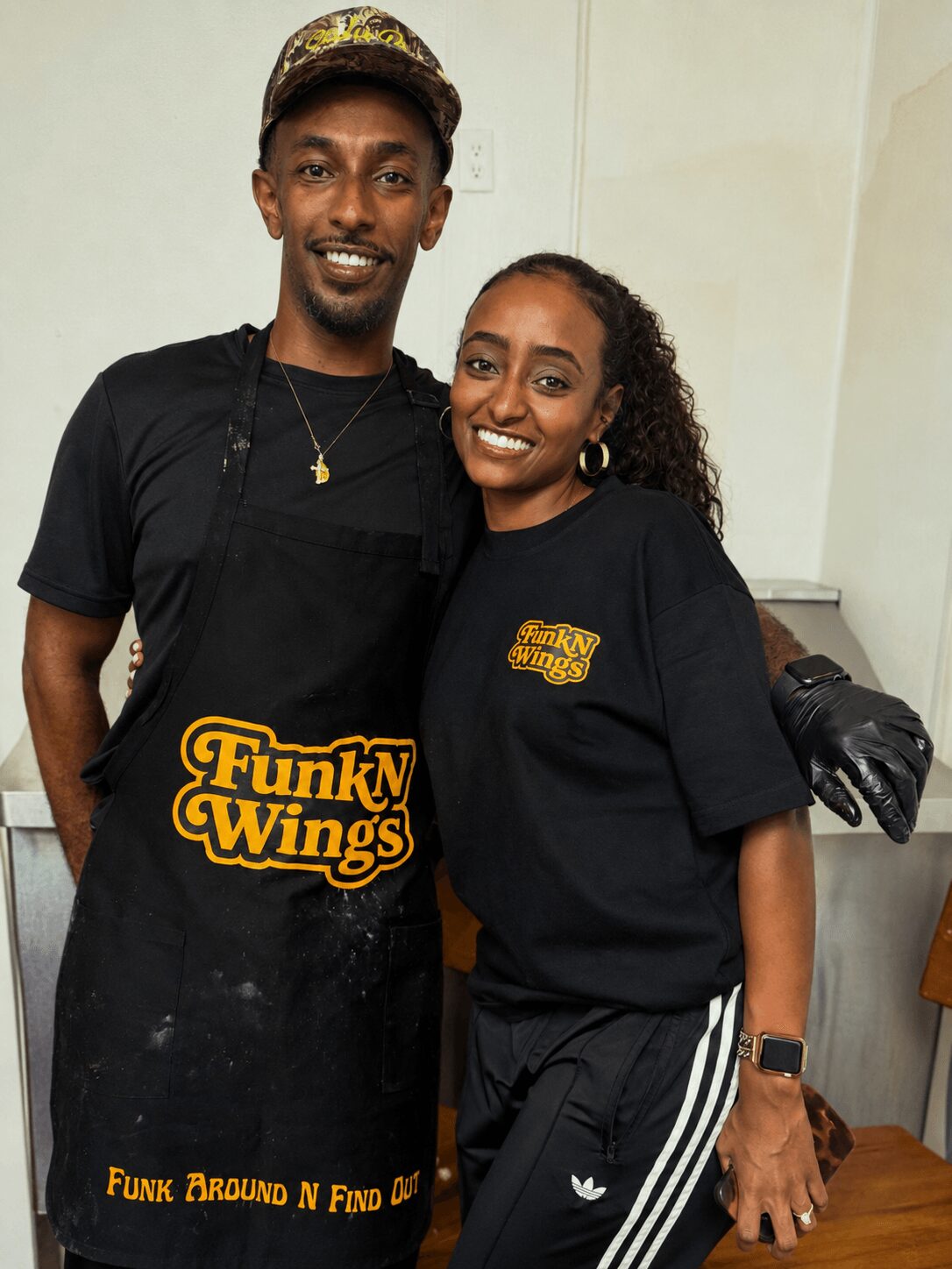 Two people smiling, standing indoors, one wearing a black apron with 'FunkN Wings' logo, the other in a black T-shirt, holding a camera.