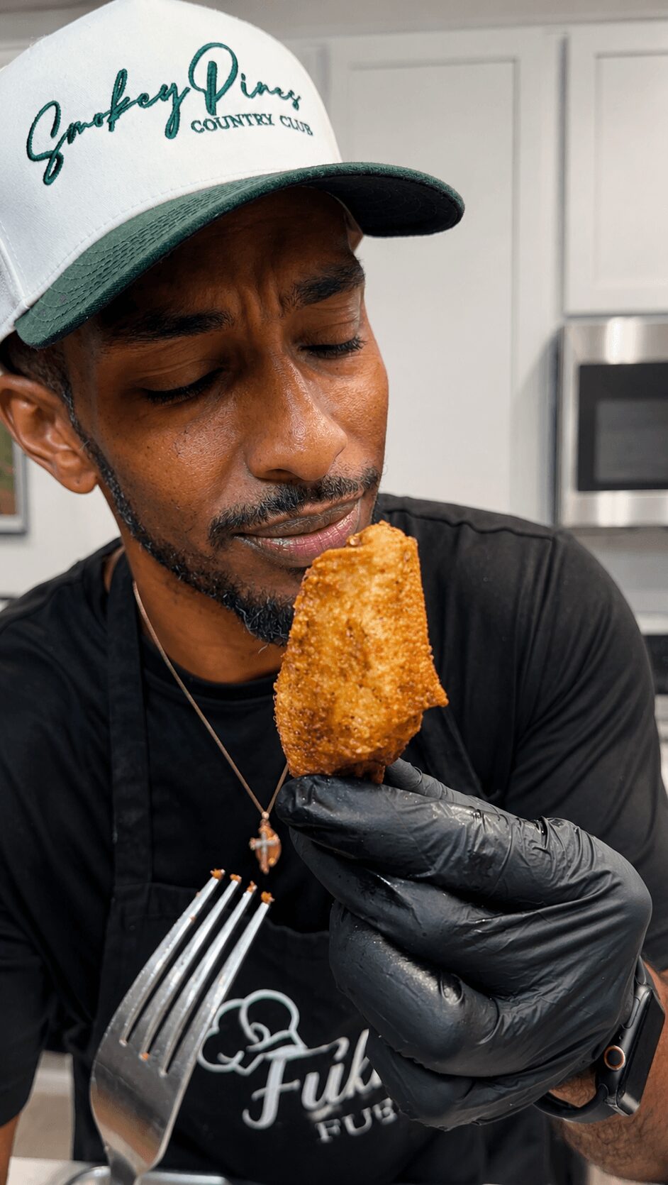 Man holding a fried food item, wearing a black glove, in a kitchen setting.