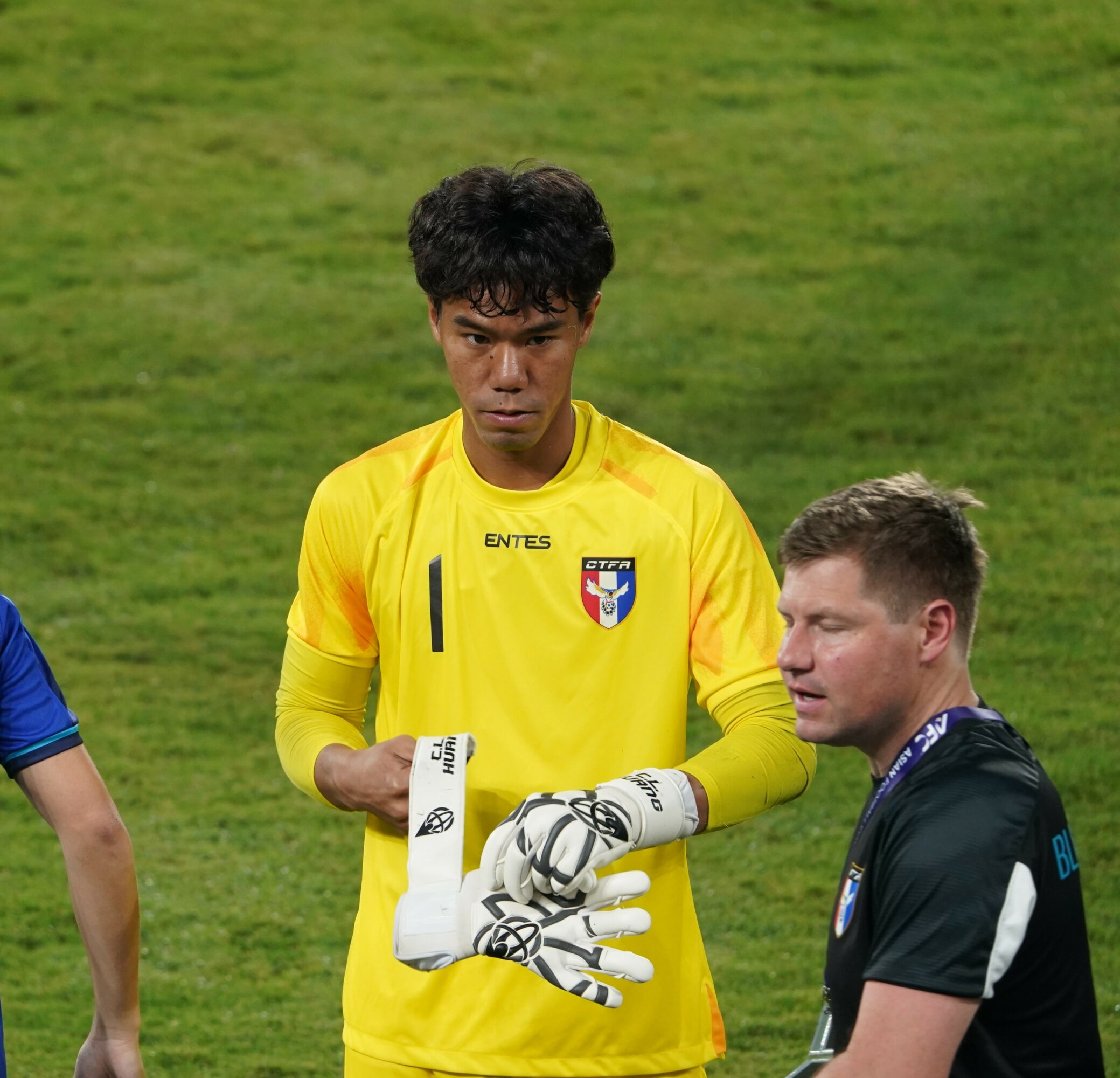 Two soccer players on the field, one in a yellow goalkeeper jersey and gloves, the other in a dark jersey, standing and talking.