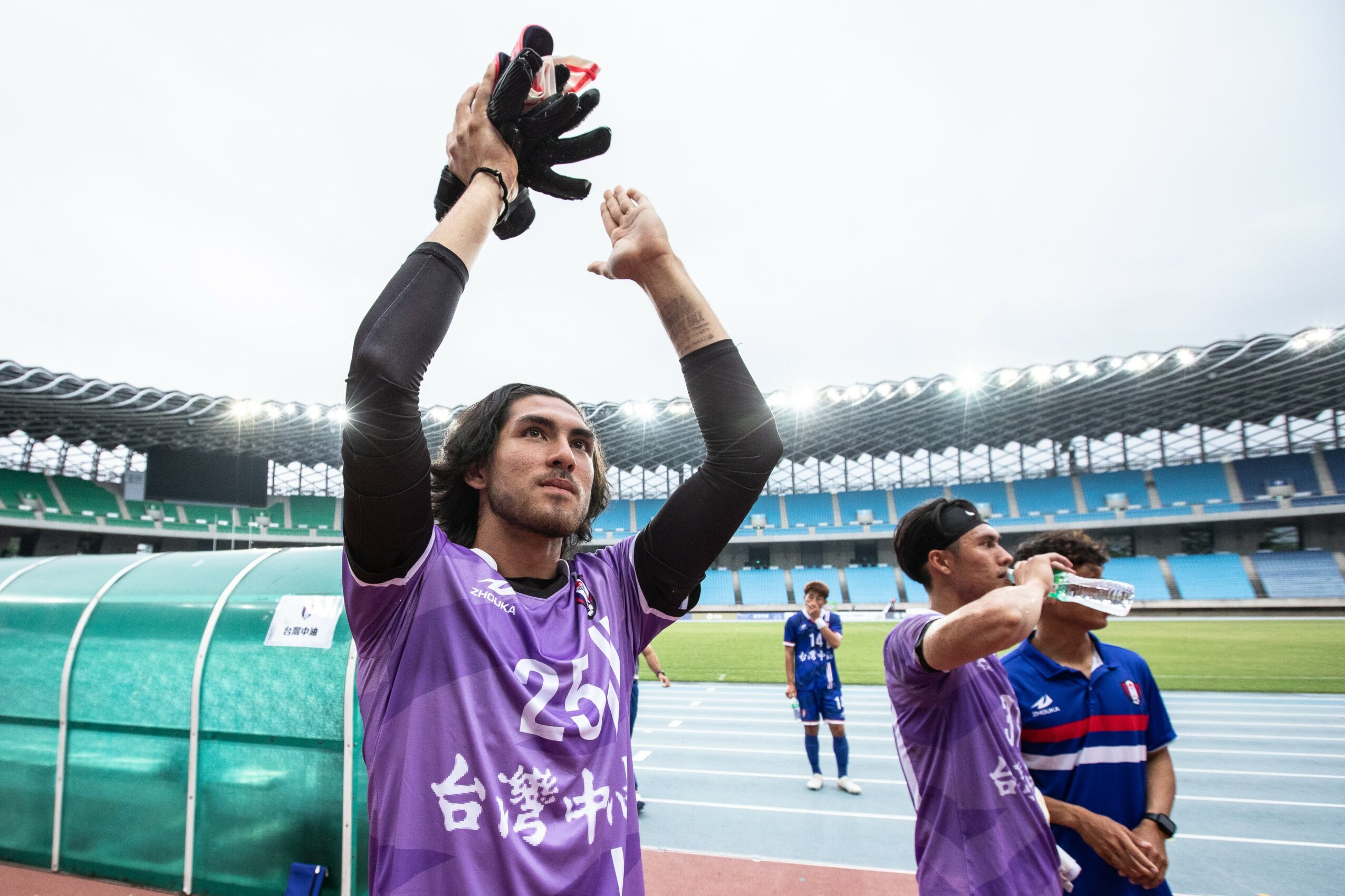 Soccer player in purple jersey raising gloved hands, standing on field with stadium and other players in background.