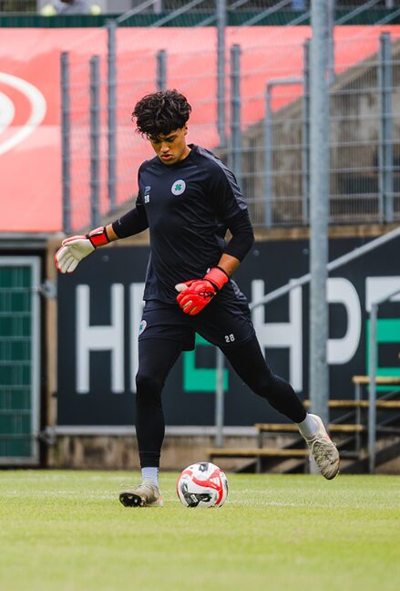 Soccer goalkeeper in black uniform and red gloves standing on grass with a ball, stadium background.