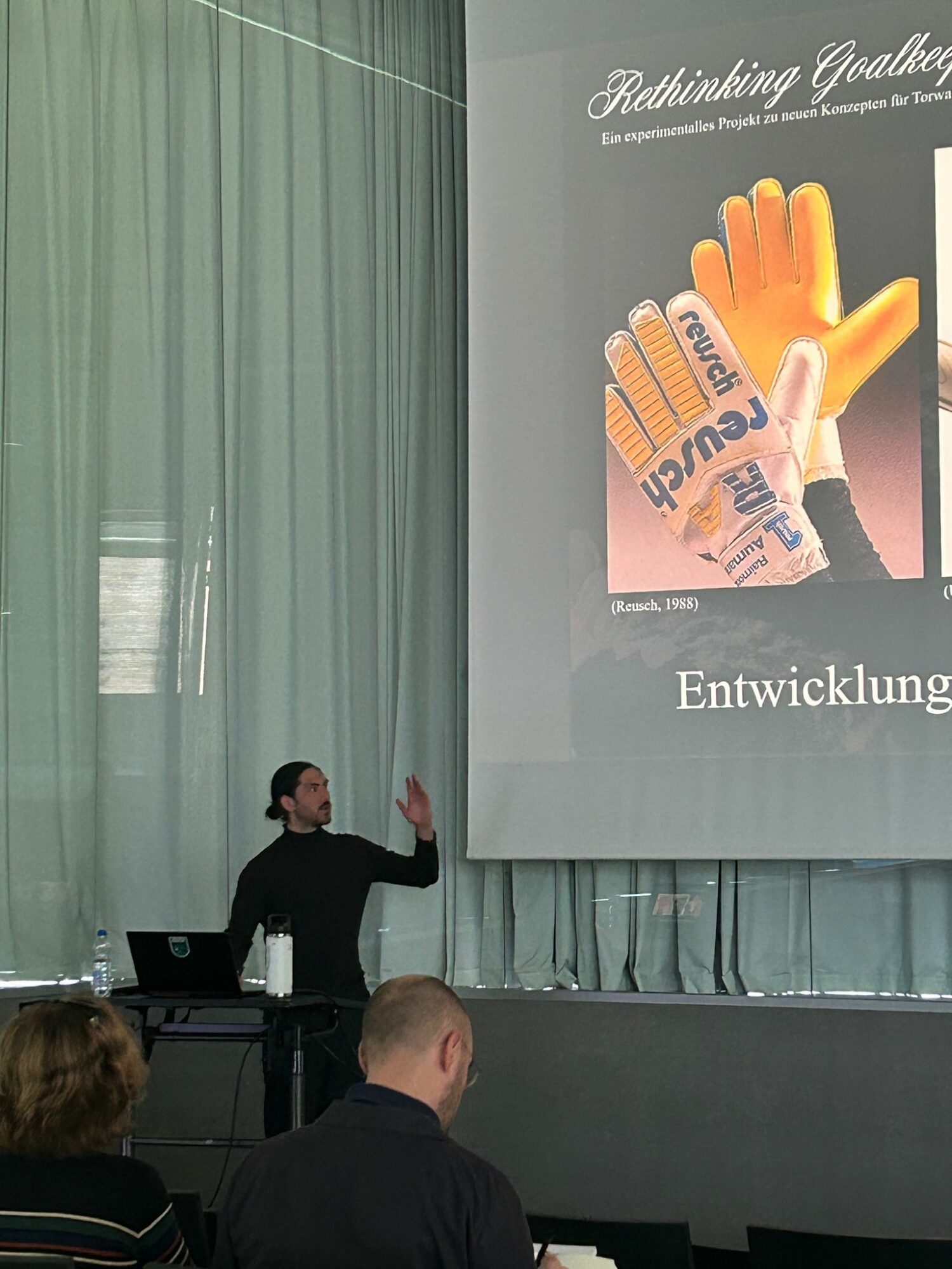 Person giving a presentation in front of a large screen with a slide showing a hand with words on fingers, in a conference room.