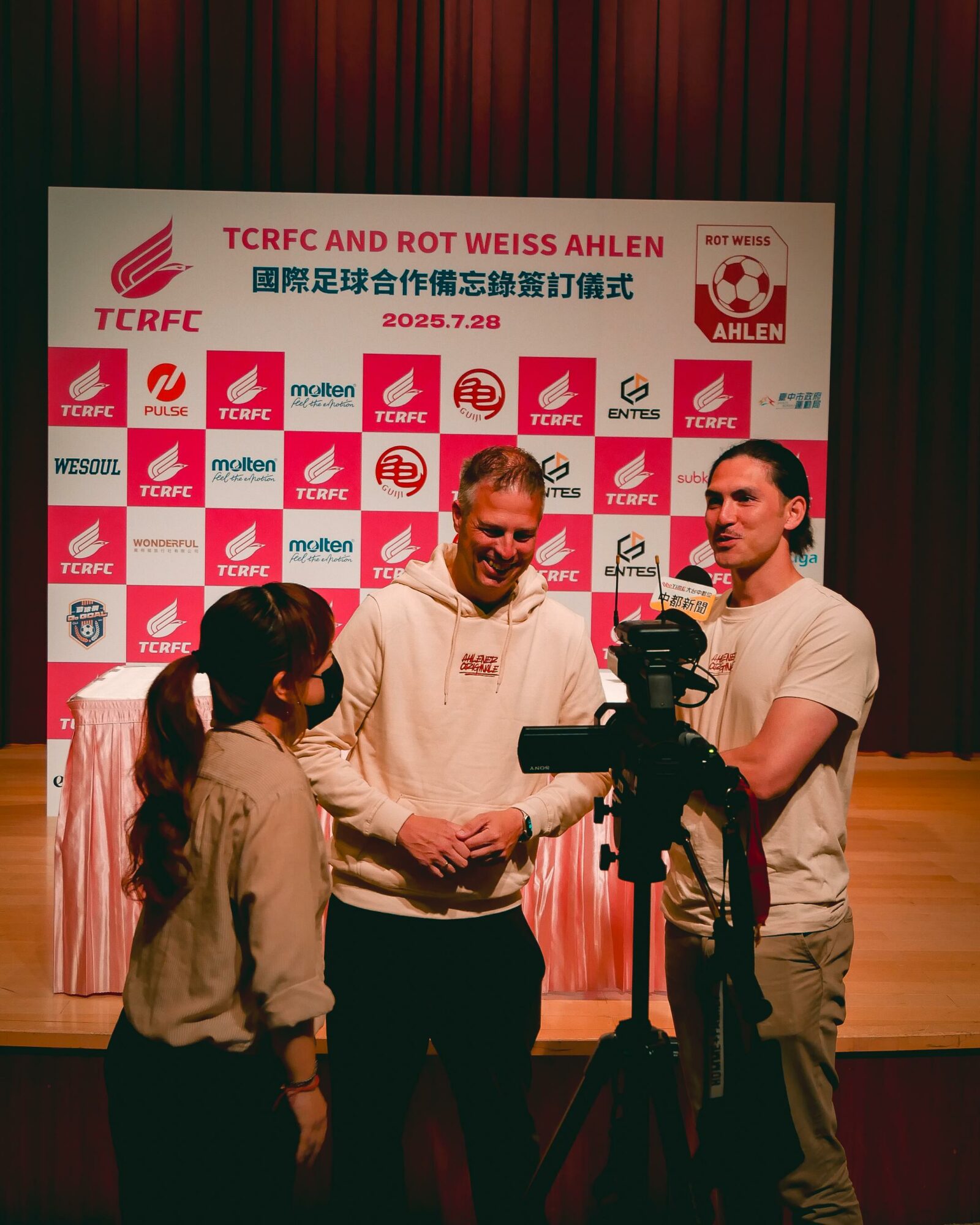 Three people on stage in front of a backdrop with logos and text, engaged in conversation, with a camera in the foreground.