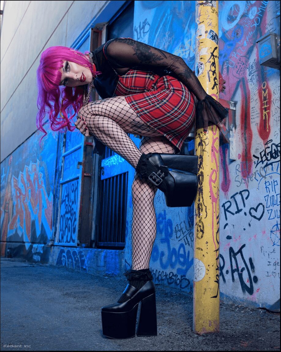 Person with pink hair in fishnet stockings and plaid skirt leaning against a yellow pole in front of graffiti wall