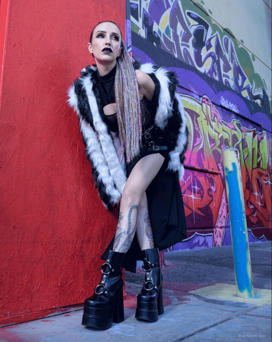 Person with long hair, makeup, and tattoos leaning against a red wall with colorful graffiti, wearing a fur coat and platform boots.