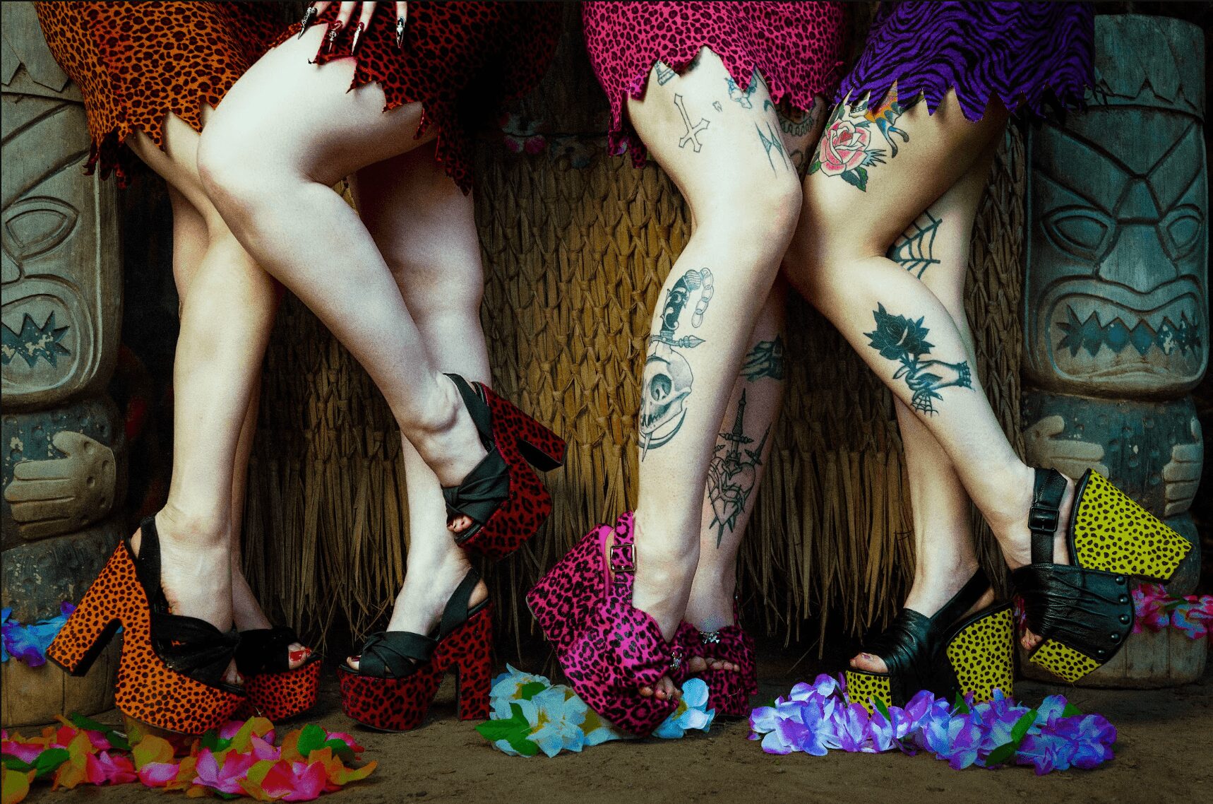 Two women with tattoos wear colorful dresses and high heels, standing with legs crossed, surrounded by scattered colorful paper and shoes.