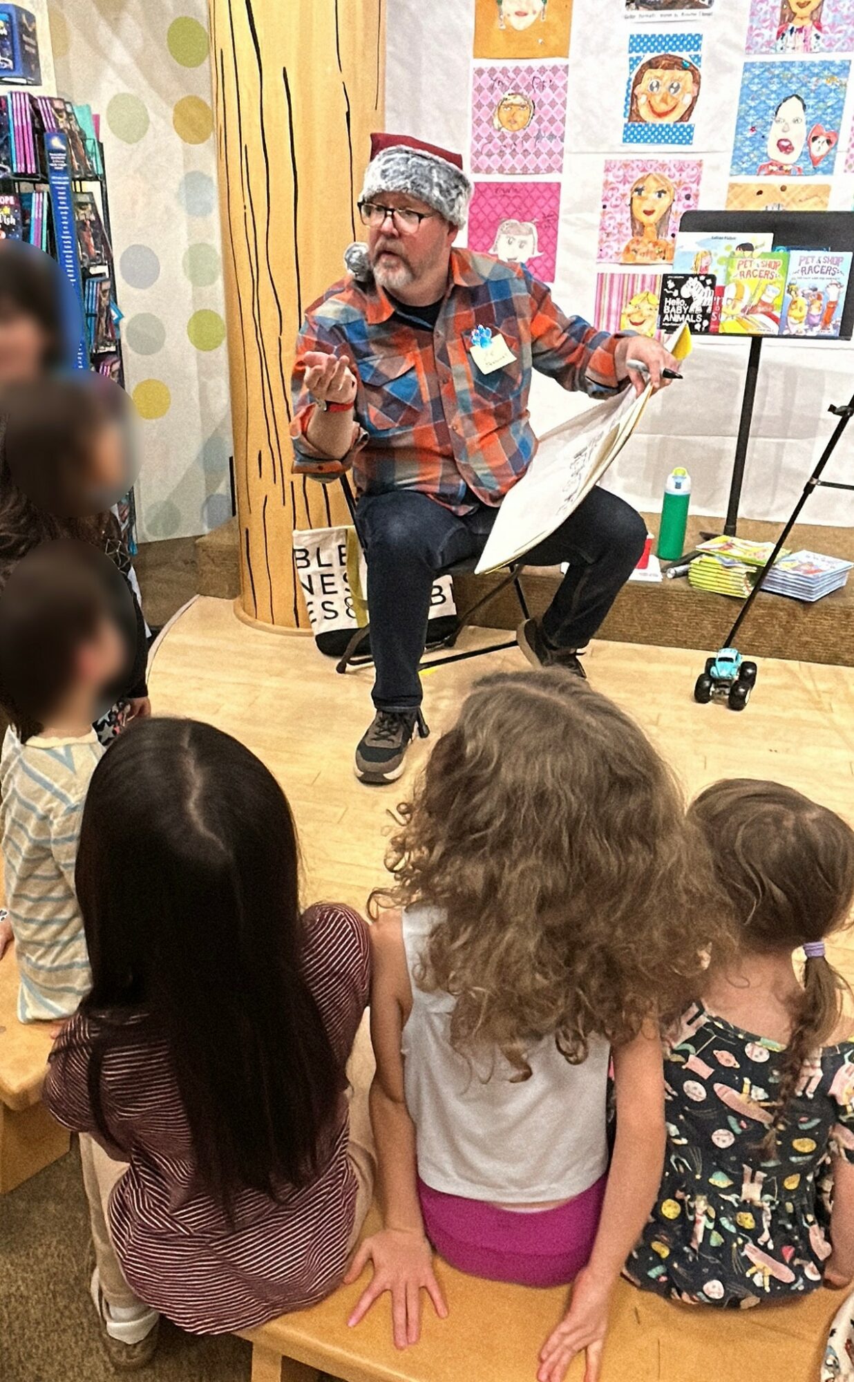 Man with glasses and a hat reads a book to children in a colorful room, with books and posters on the wall.