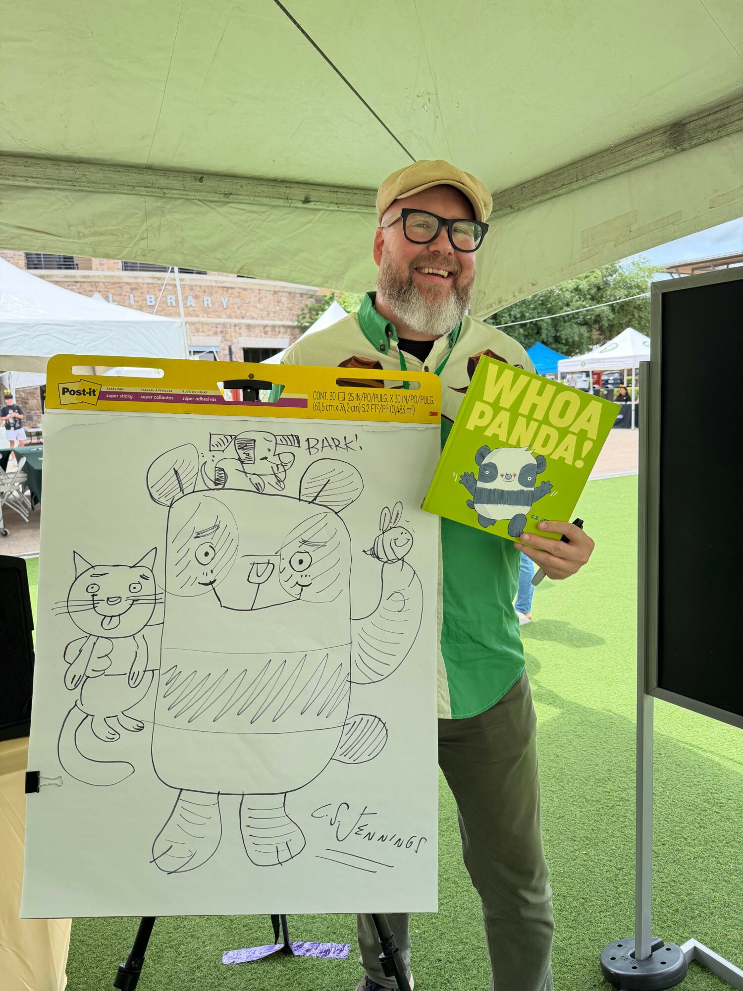 Man with glasses and a beard holding a birthday card and a large drawing of a dog with three smaller animals, under a tent.