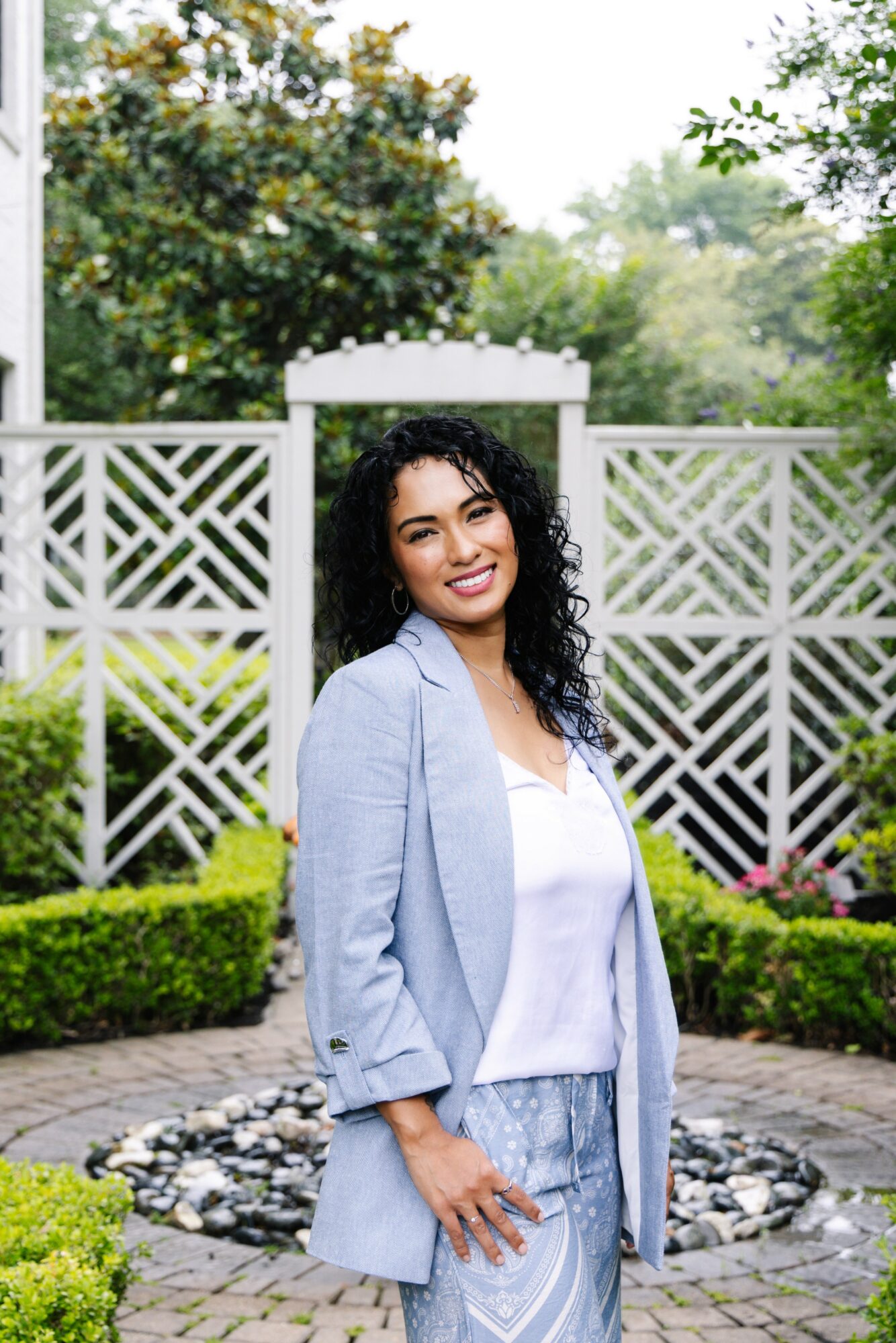 Woman with curly hair smiling outdoors, standing in front of a white lattice fence and greenery.