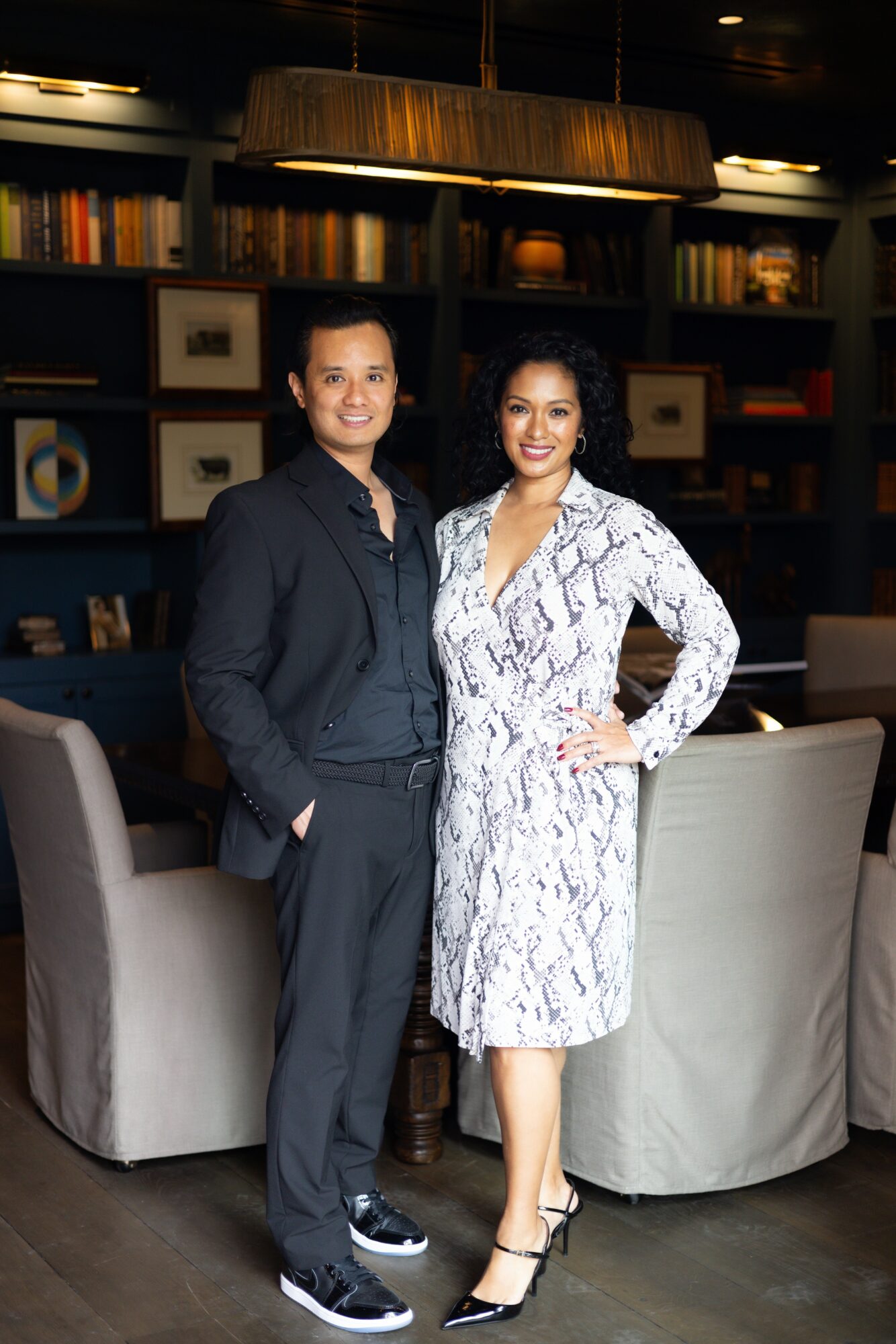 Two people standing in a room with bookshelves and chairs, smiling at the camera.