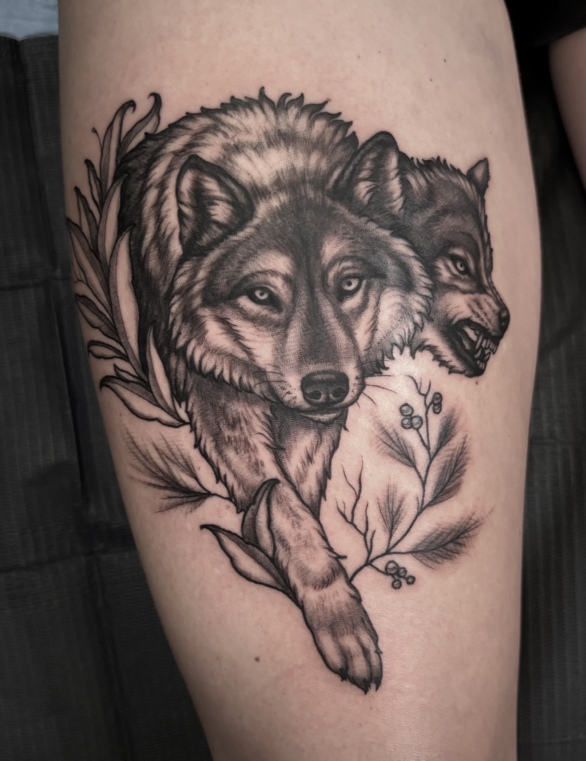 Tattoo of a wolf and a dog with detailed fur, surrounded by leaves and small plants, in black and gray ink.