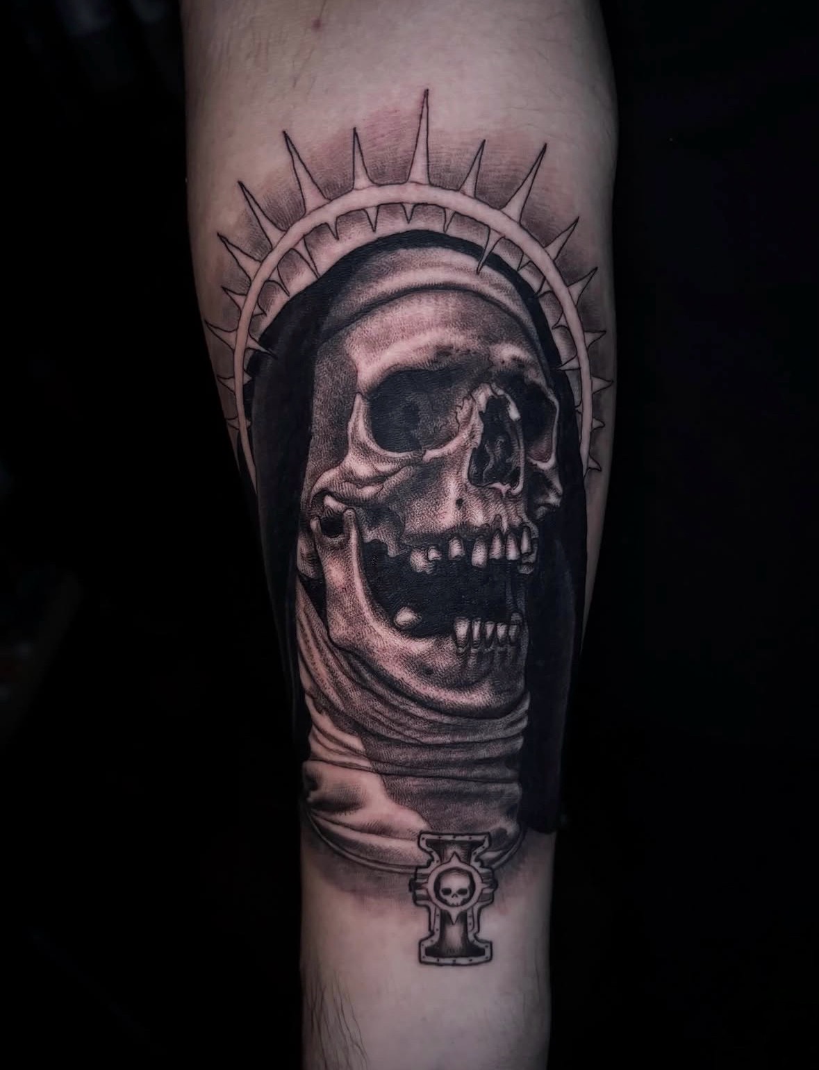 Tattoo of a skull with a halo and a small symbol below, in black and gray shading.