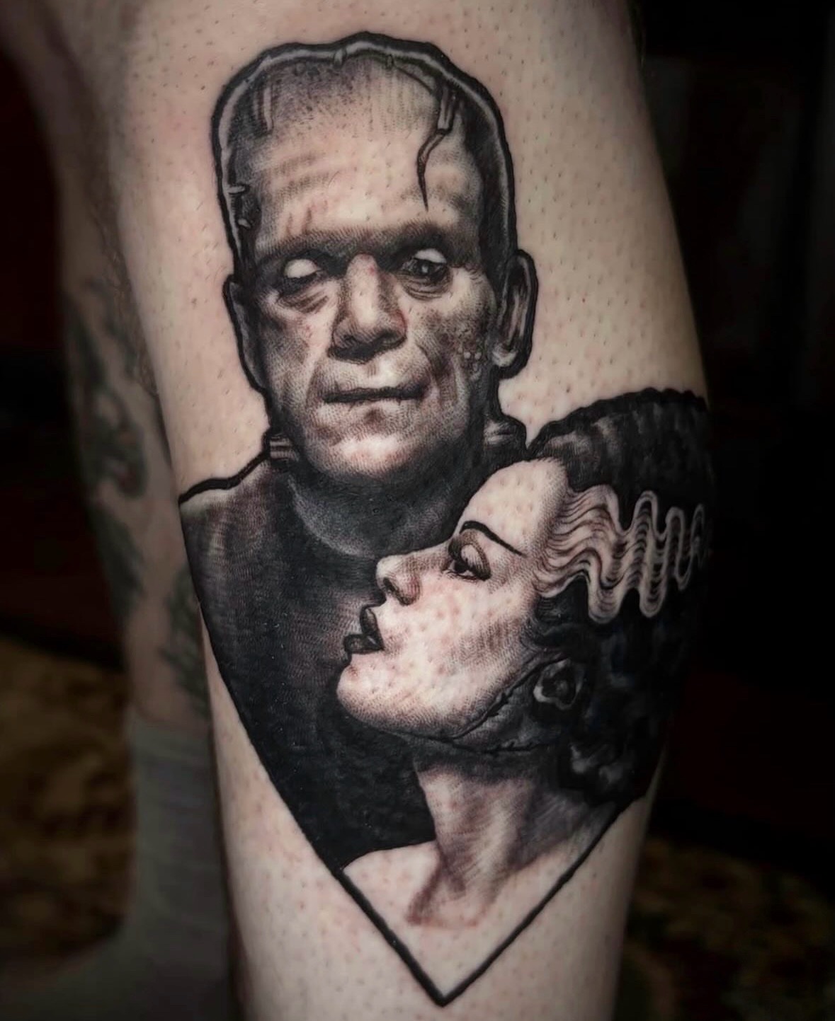 Black and gray tattoo of Frankenstein's monster and a woman with curly hair, facing each other, on skin.
