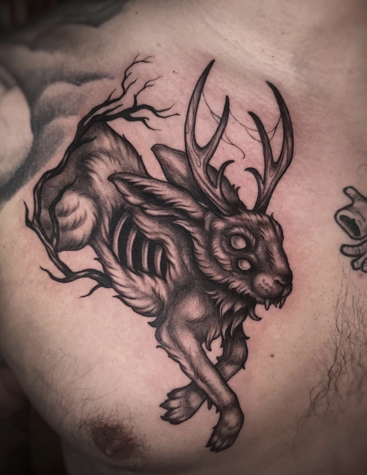 Tattoo of a rabbit with antlers, detailed shading, and a tree branch on its back, on a person's chest.