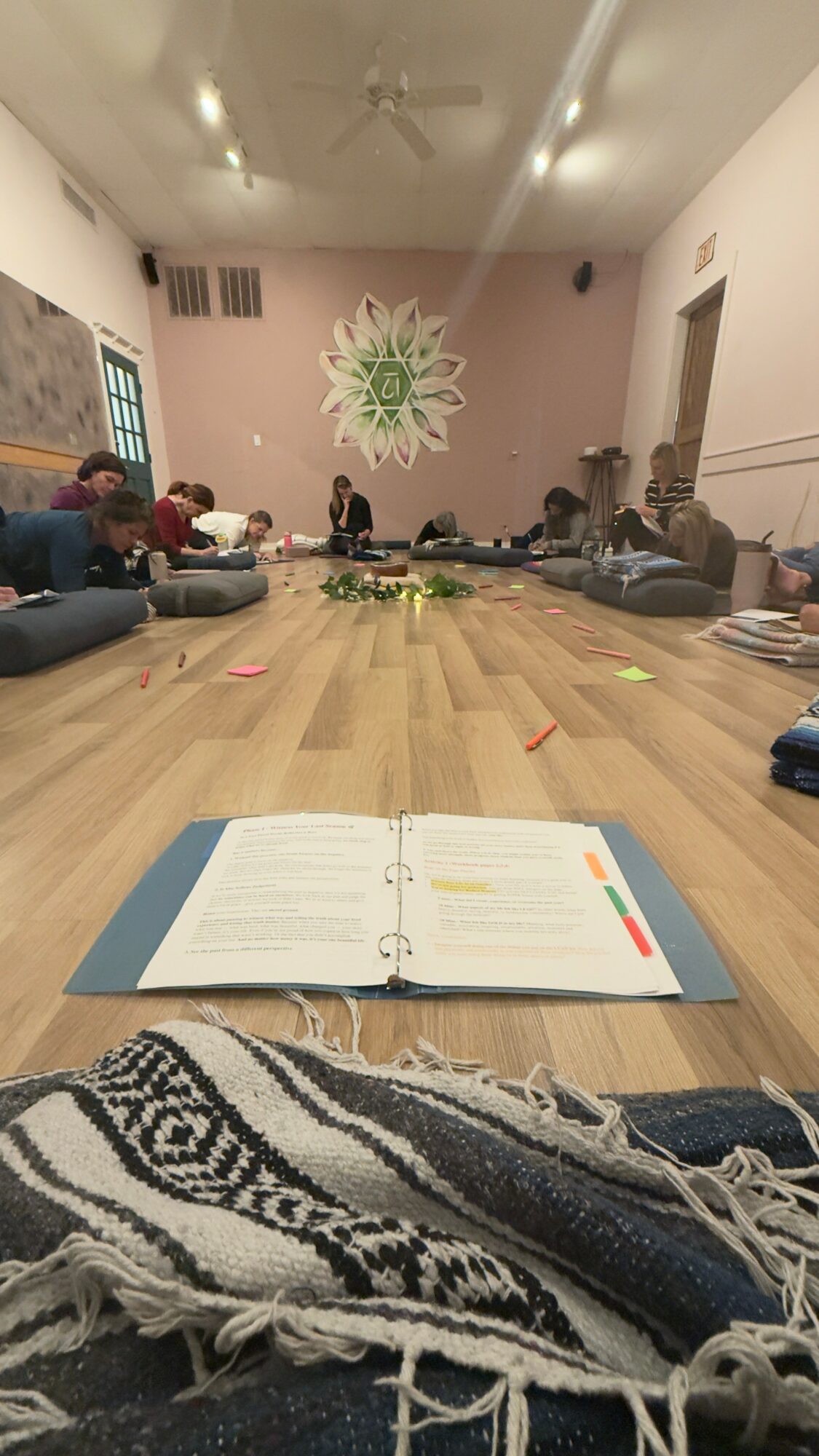 People sitting around a large table in a room with a decorative wall art, open notebook in foreground, and a patterned cloth.