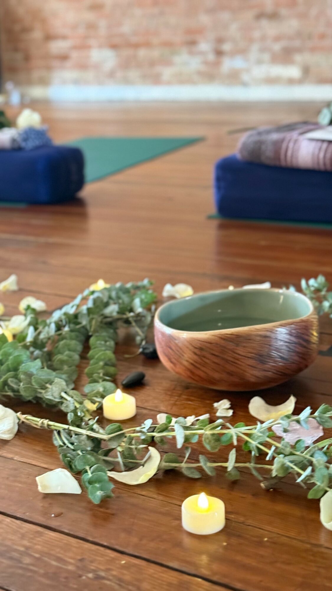 Decorative bowl surrounded by candles and greenery on wooden floor, with yoga mats and brick wall in background.