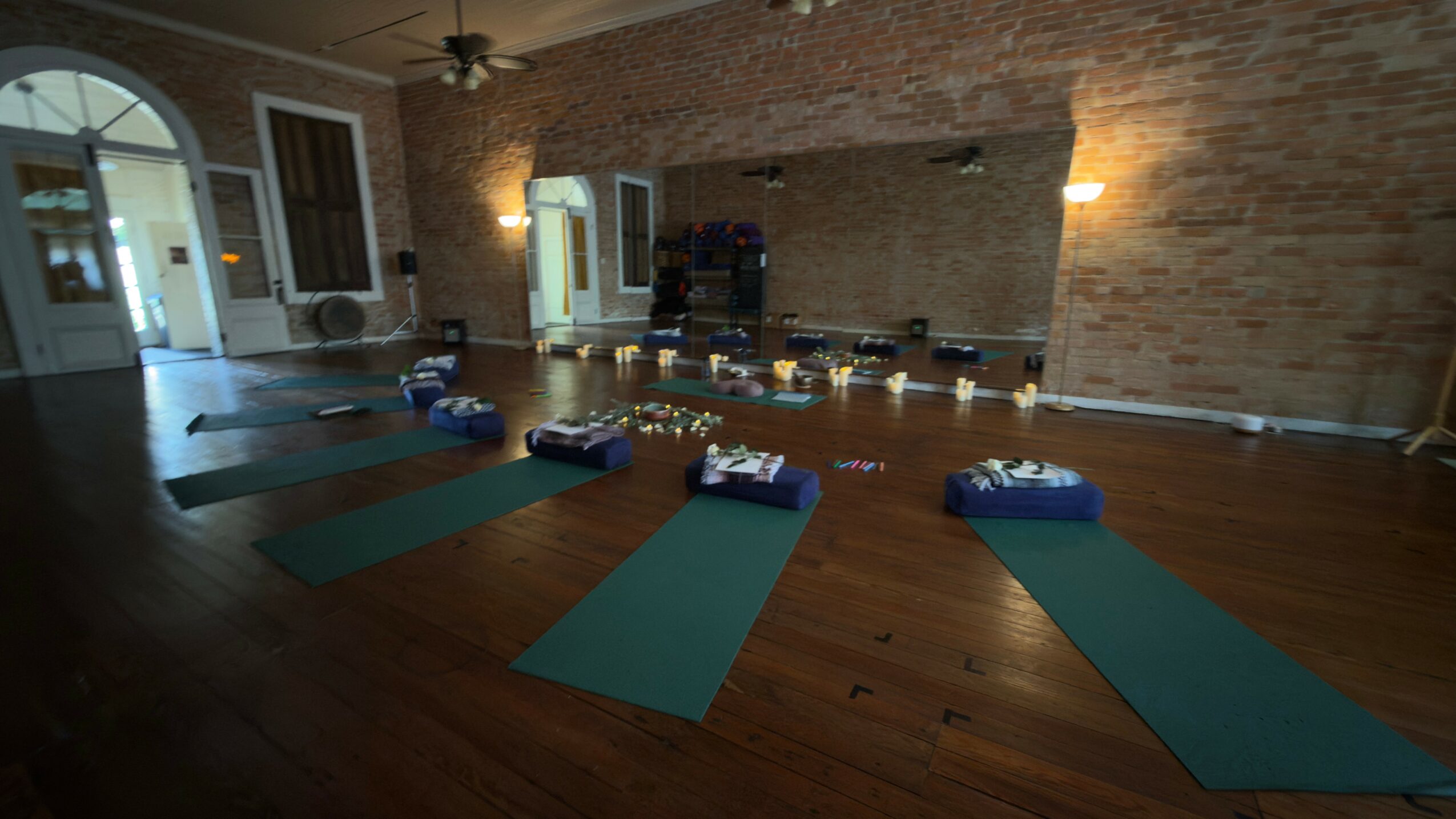 Yoga mats and props arranged in a studio with brick wall and wooden floor, windows, and lighting.