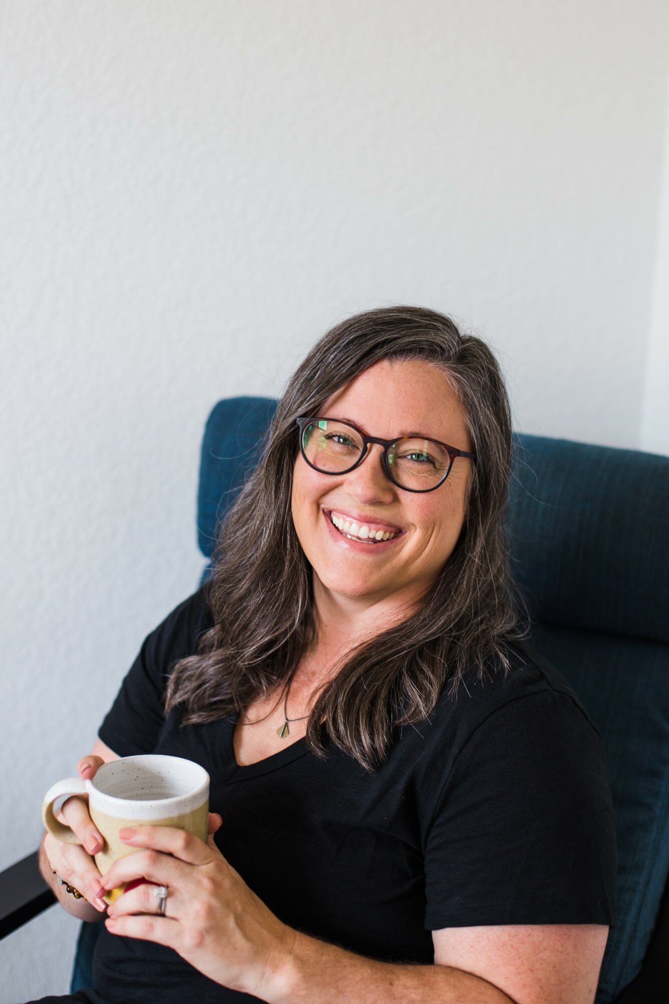 Woman with glasses smiling, holding a mug, sitting on a blue chair against a plain white wall.