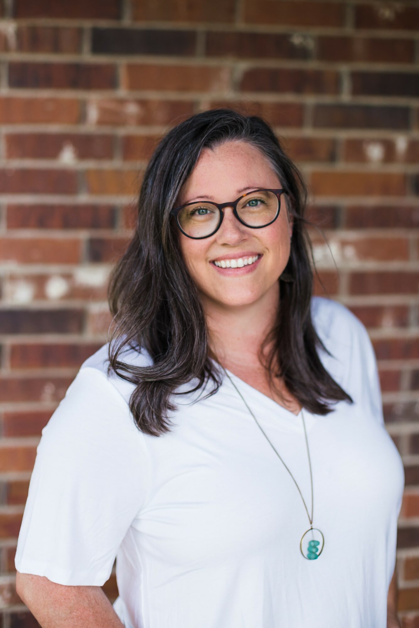 Smiling woman with glasses and dark hair, wearing a white top, standing in front of a brick wall.