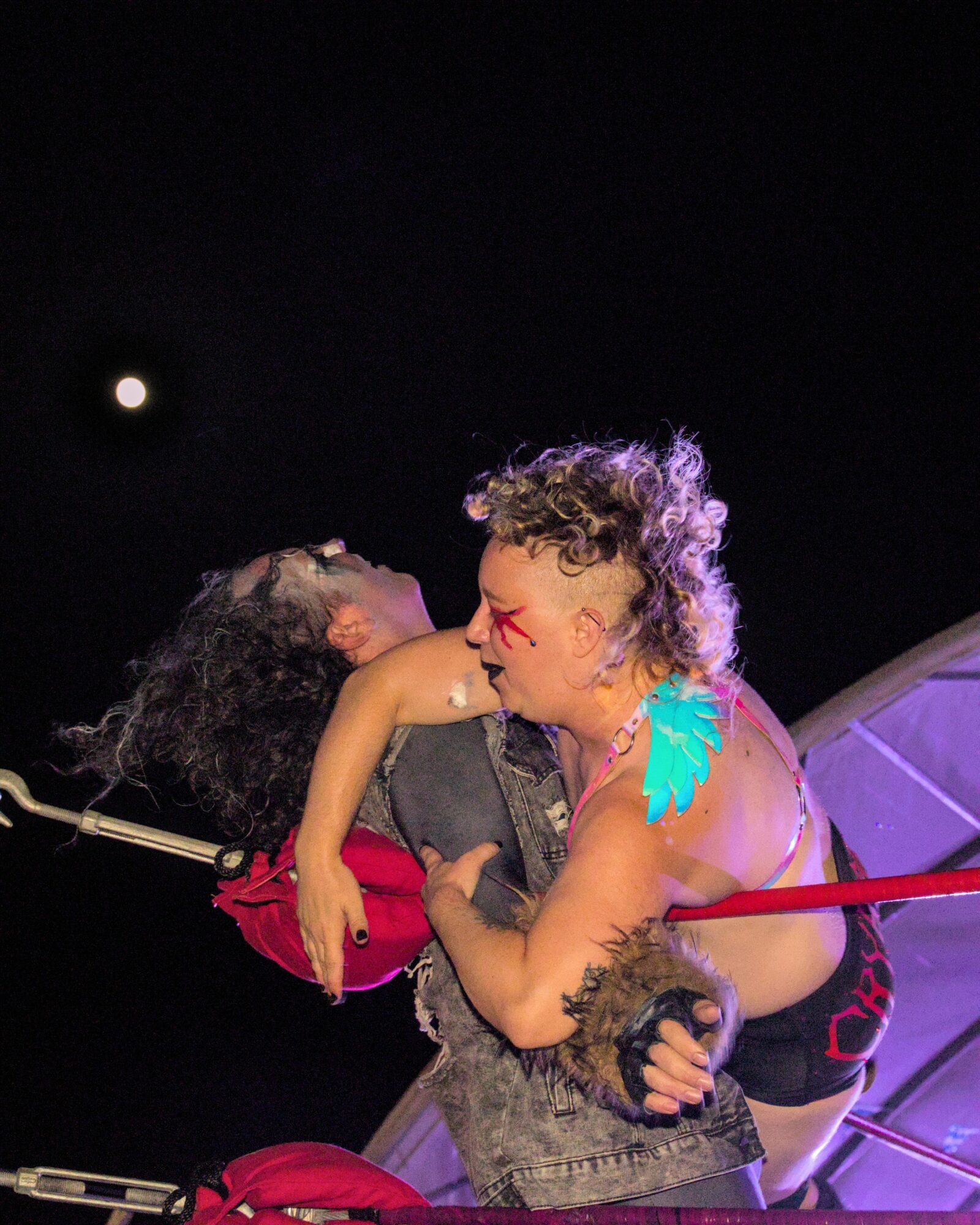 Two people in costume perform on stage with a dark background and a bright moon, one with curly hair and makeup.