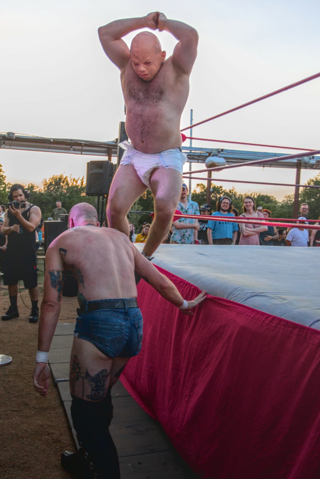 Two men preparing for a wrestling match outdoors, one standing on the ring edge, the other reaching up, crowd watching.