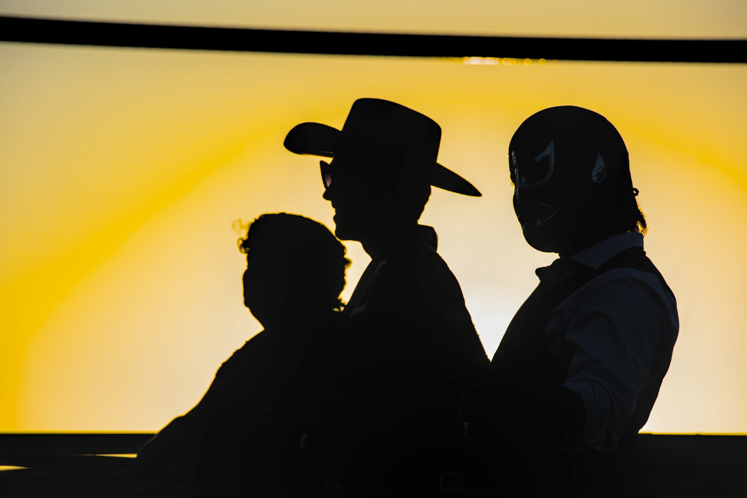 Silhouettes of three people sitting against a yellow-orange background, one wearing a hat, another with a mask, in profile view.