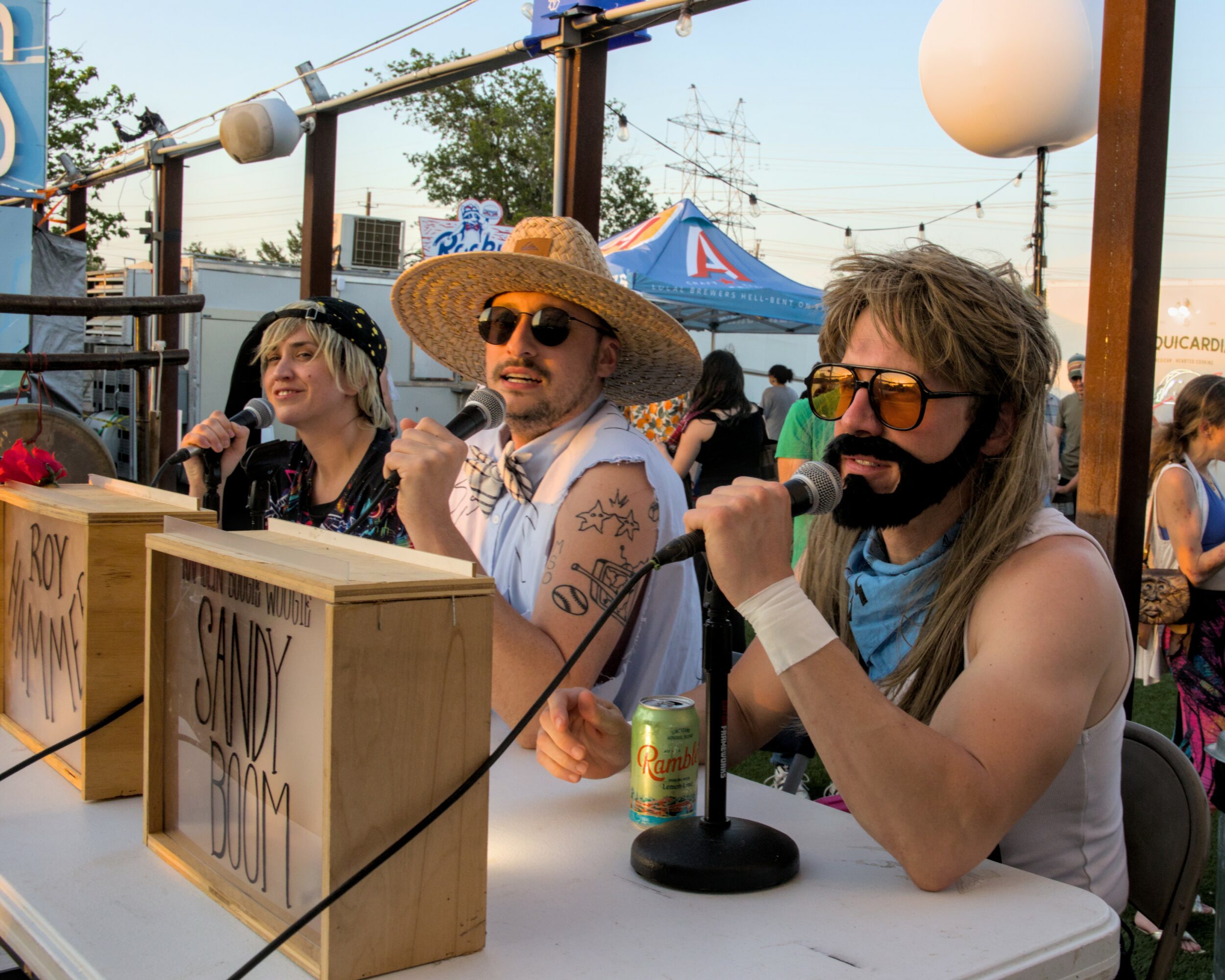 Three people sitting at a table outdoors, with boxes and a microphone, during a sunny event.