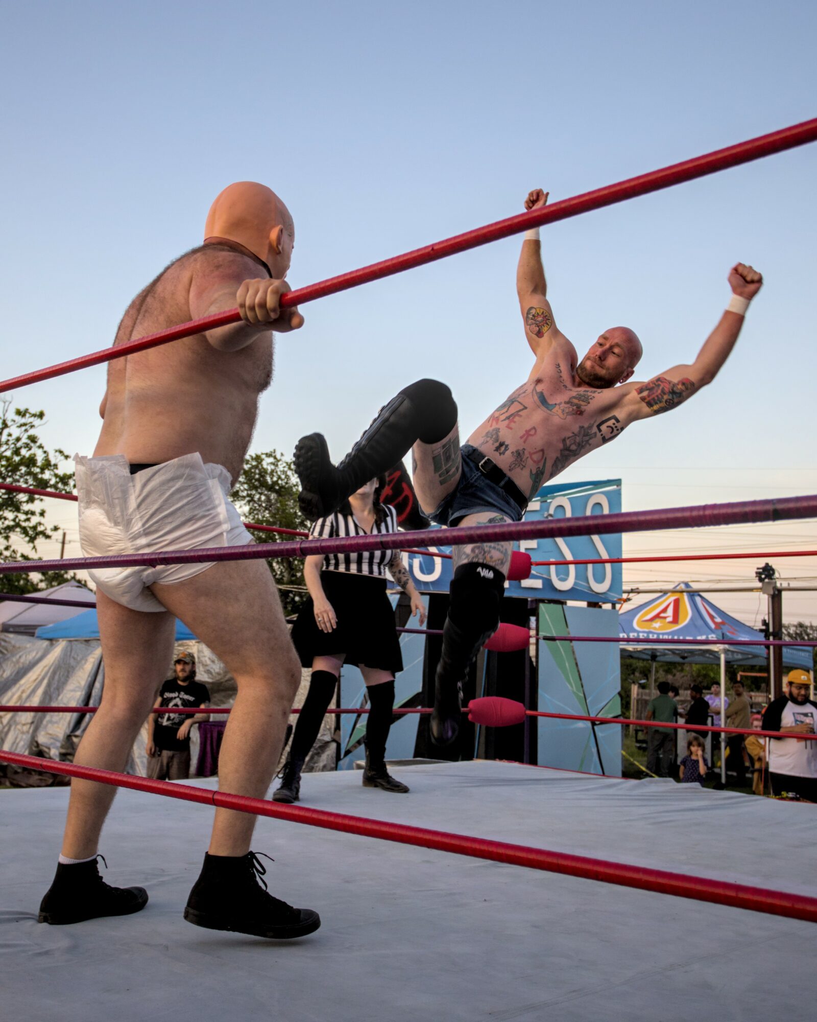 Two men wrestling in a ring outdoors, one with tattoos and the other bald, with a crowd and carnival rides in background.