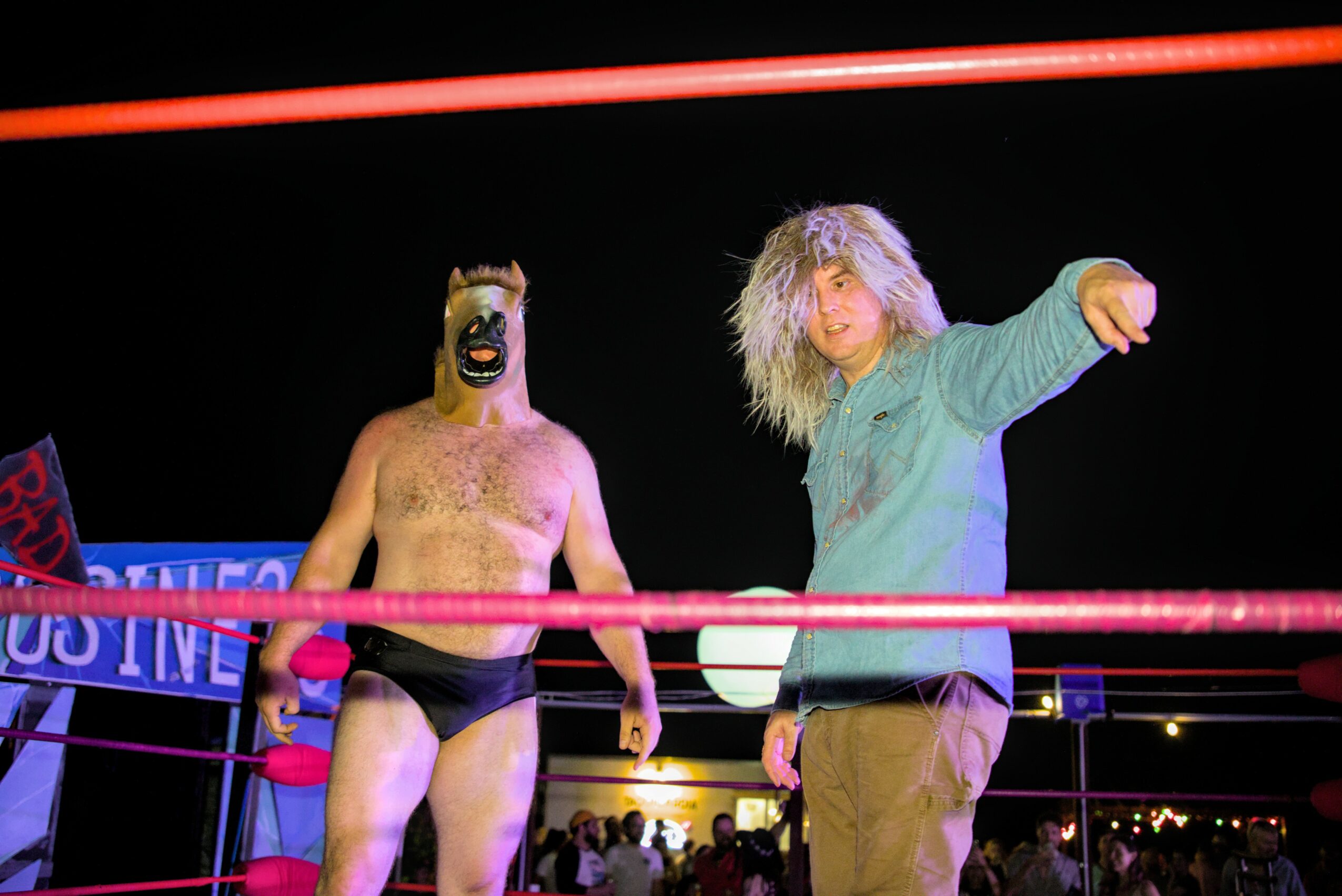 Person wearing a mask and black shorts in a wrestling ring, with another person pointing outside the ring, crowd in background.