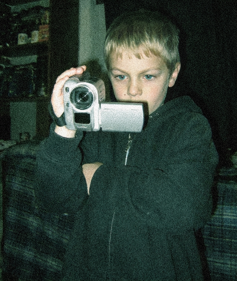 Boy with blond hair holding a video camera, wearing a dark hoodie, in a dimly lit room.