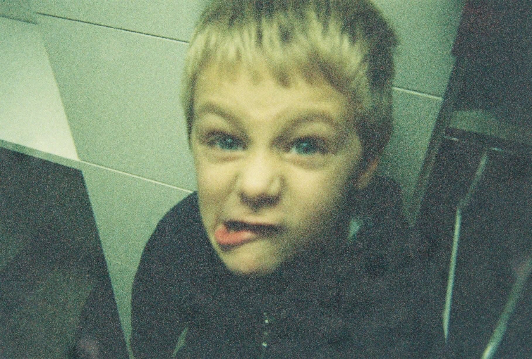 Child making a funny face with tongue out, in a close-up shot, with tiled wall background.