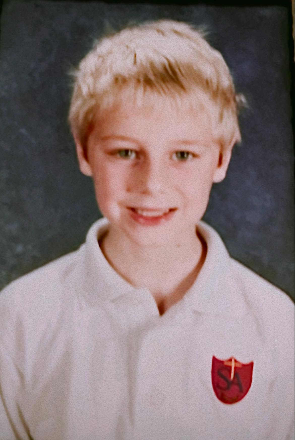 Young boy with blonde hair wearing a white shirt with a red and gold crest, smiling, dark background.