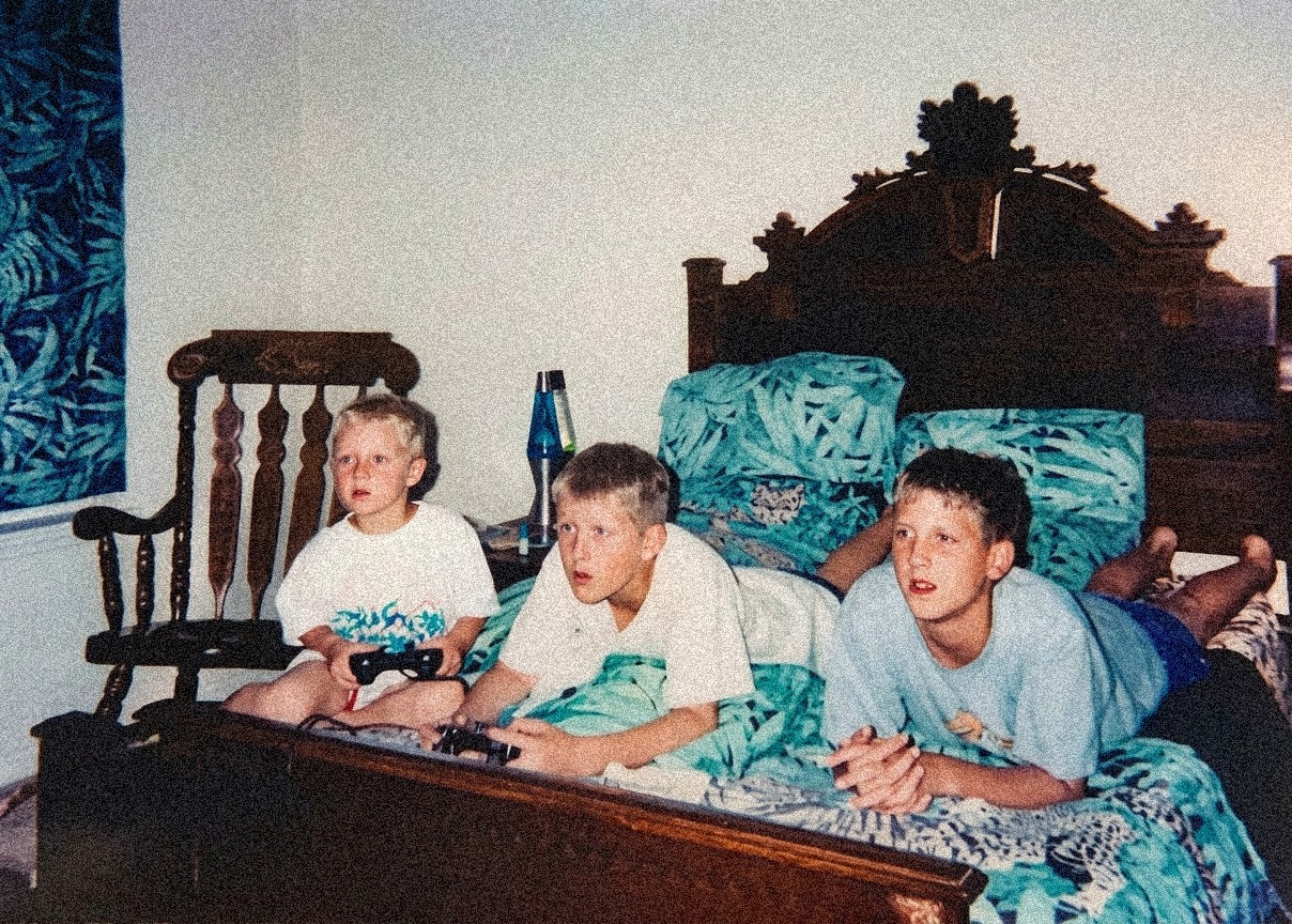 Three children lie on a bed watching television, with a wooden headboard and blue bedding, in a room with a chair and window.