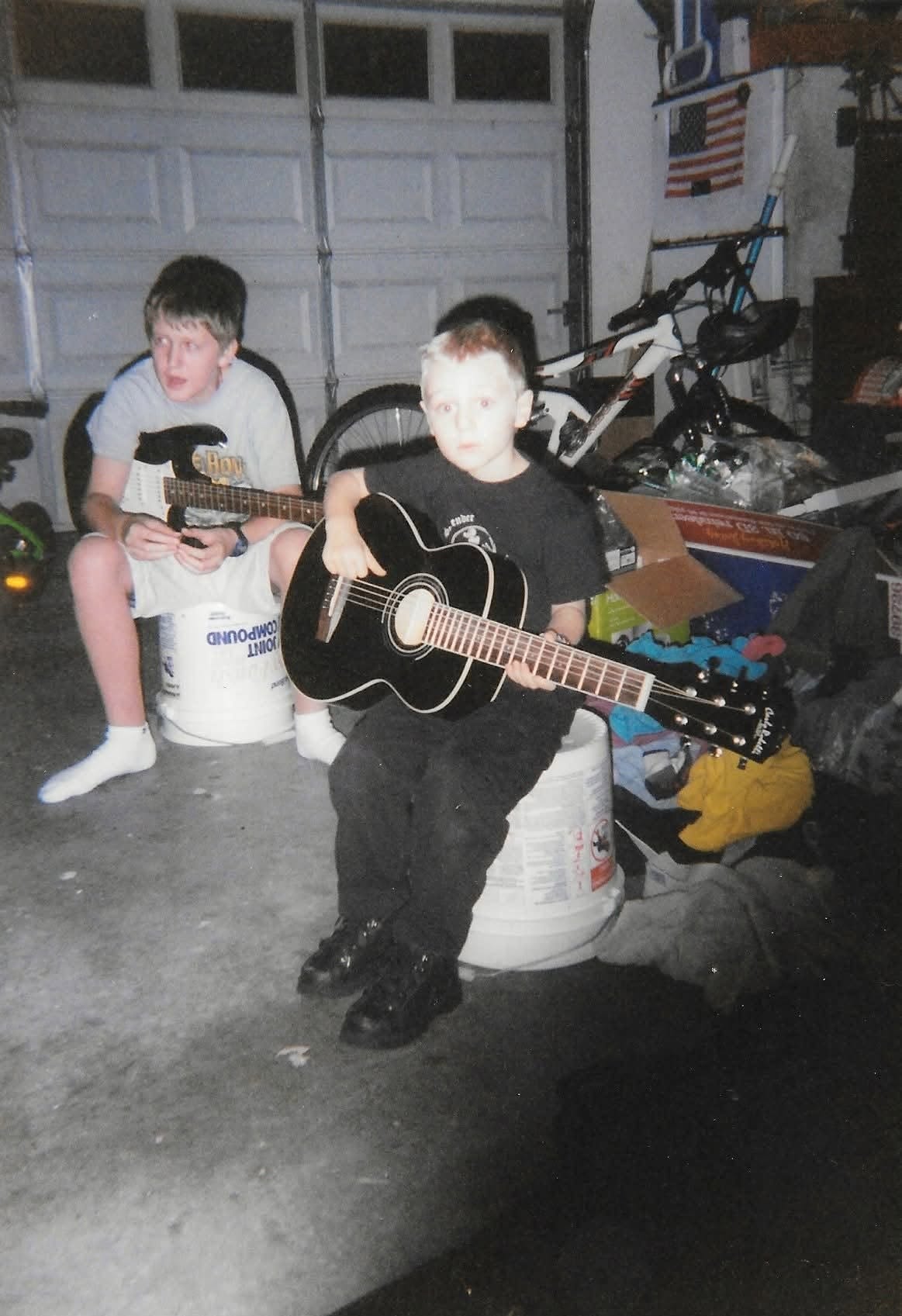 Two children, one sitting on a container and the other standing, playing guitars in a garage with bicycles and tools visible.