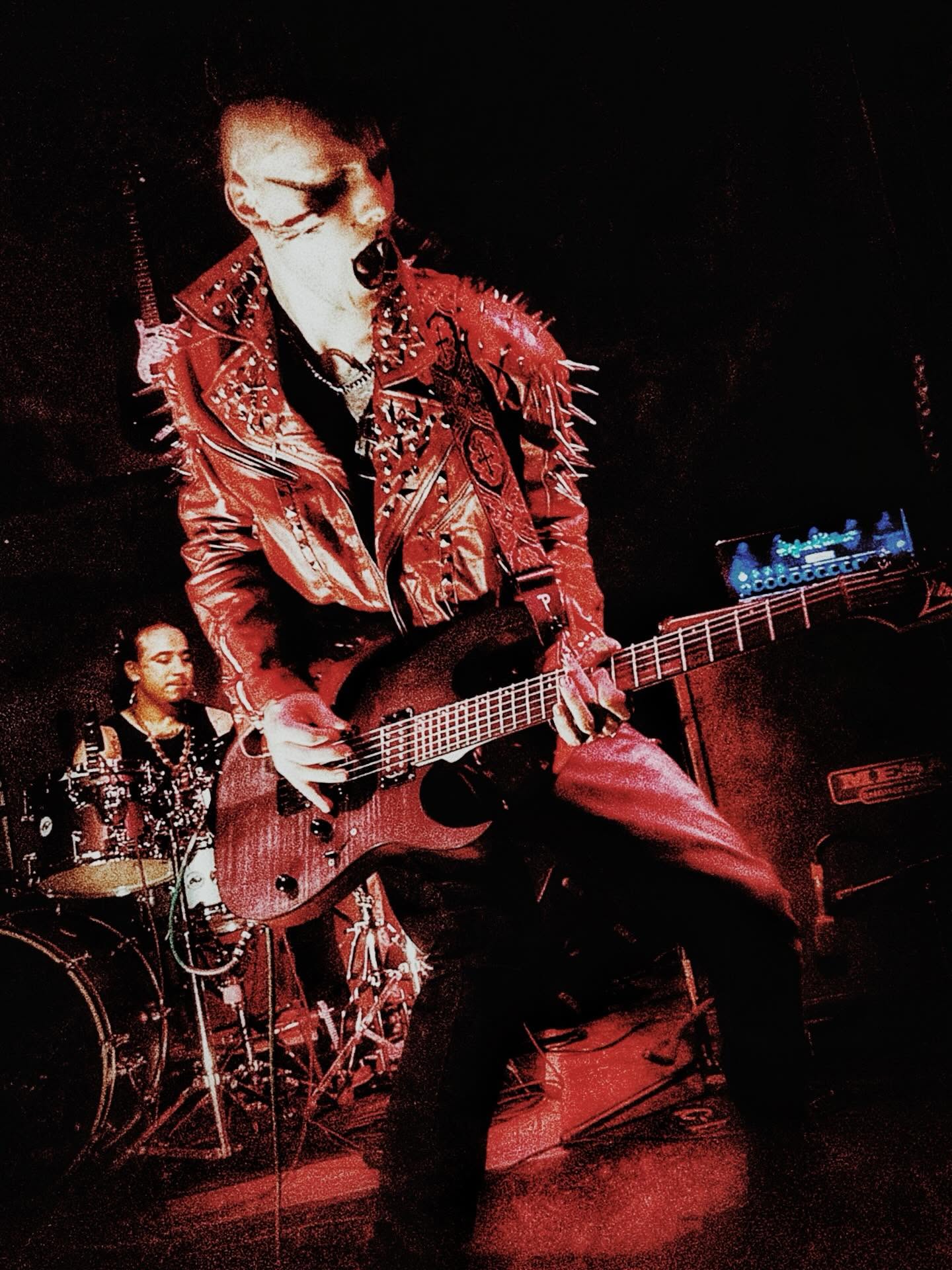 Musicians perform on stage with dark background, one playing guitar and another on drums, both dressed in dark clothing.