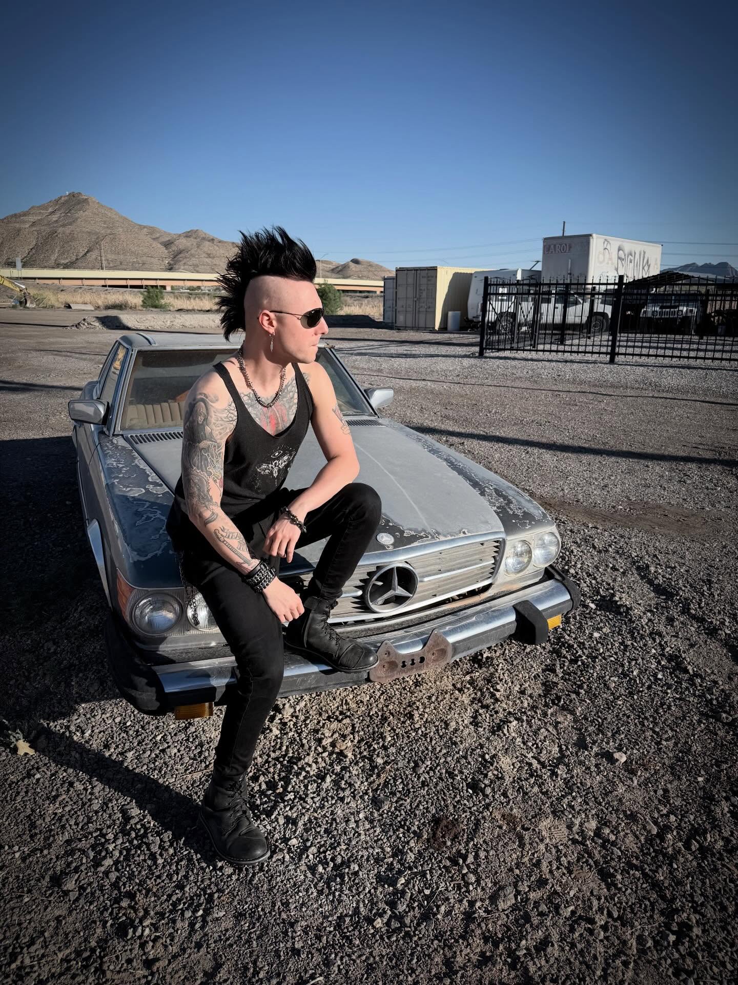 Person with tattoos and sunglasses sitting on a vintage car in a desert landscape with mountains and buildings in the background.