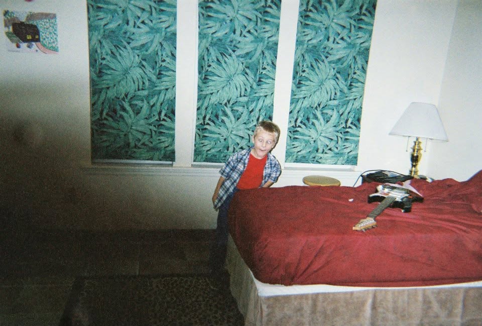 Child standing near a bed with a red cover, a lamp, and a window with leafy patterned curtains.