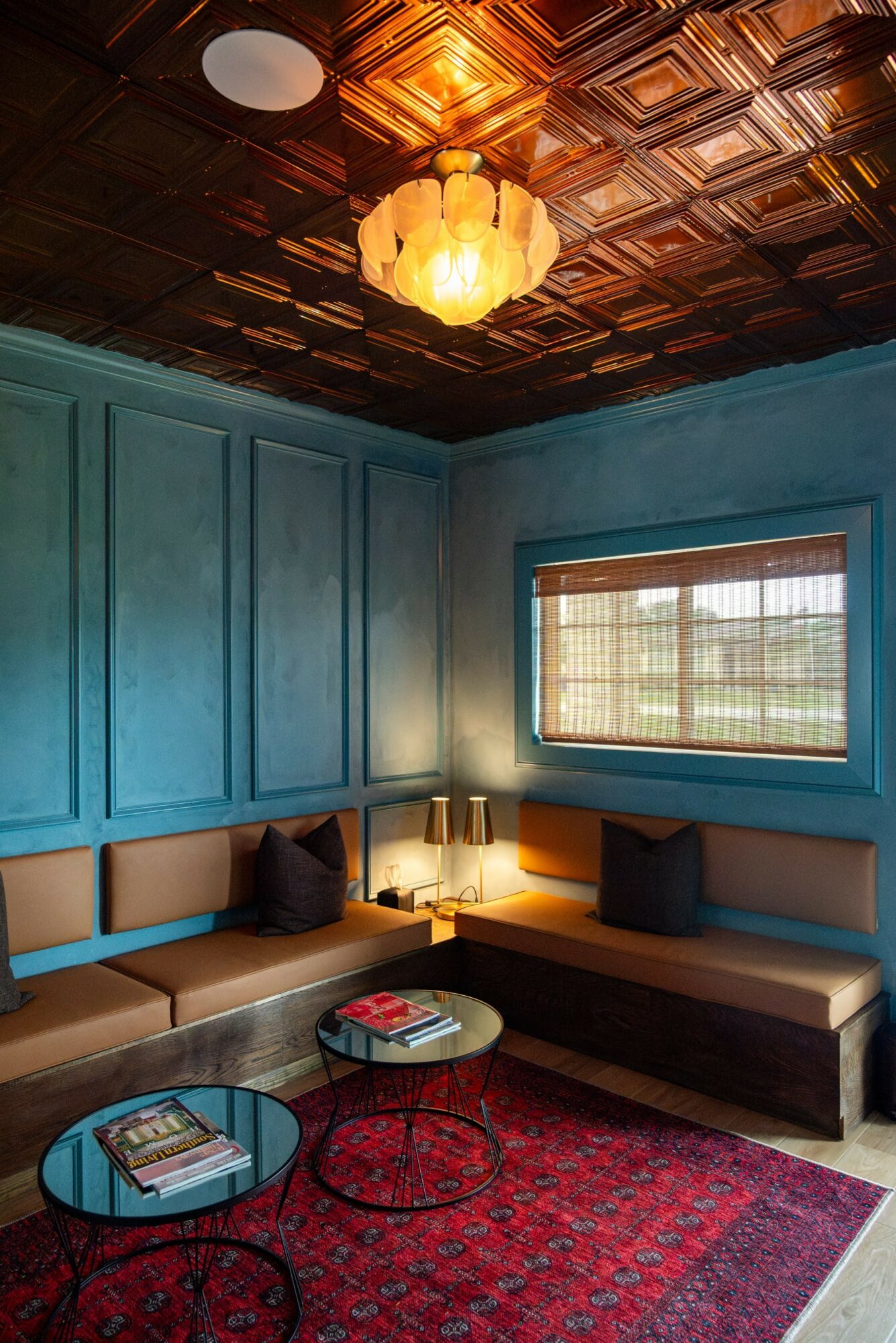 Living room with blue paneled walls, window, built-in seating with cushions, two small round tables, and a red patterned rug.