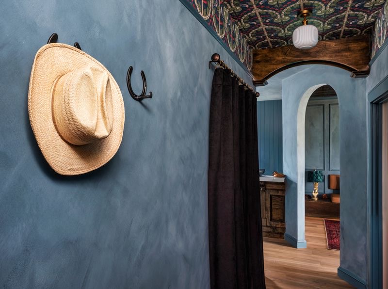 Blue wall with a straw hat and black hook, doorway with black curtain, and view into a room with wooden furniture.