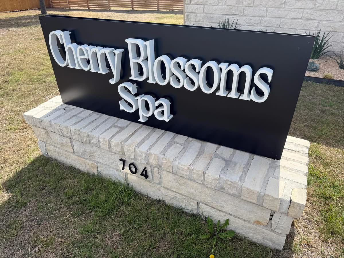 Sign for Cherry Blossoms Spa on a brick base, with a black background and white 3D text, outdoors.