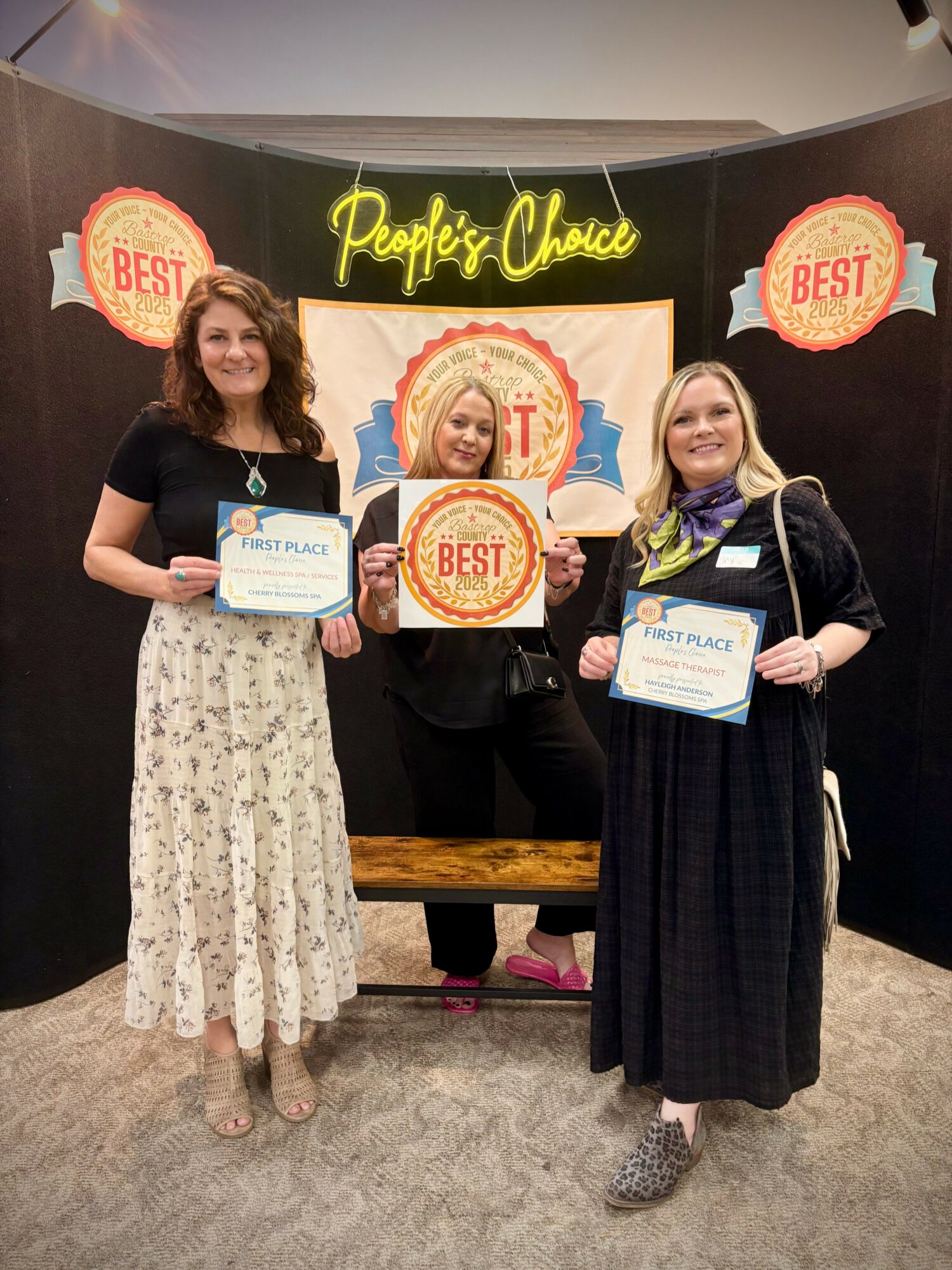 Three women holding awards and certificates in front of a backdrop with 'People's Choice' and 'Best' signs, smiling.