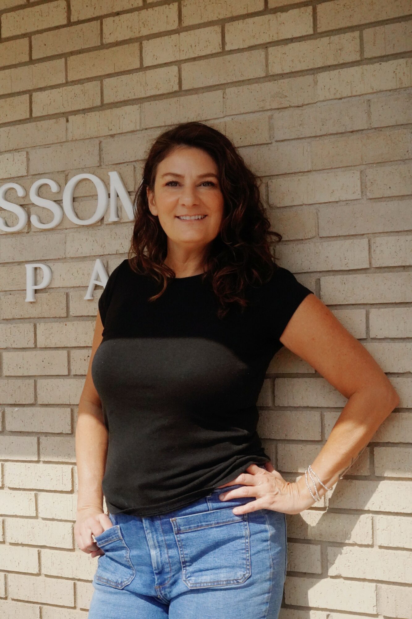 Woman with curly hair smiling, wearing a black top and blue jeans, standing against a brick wall.