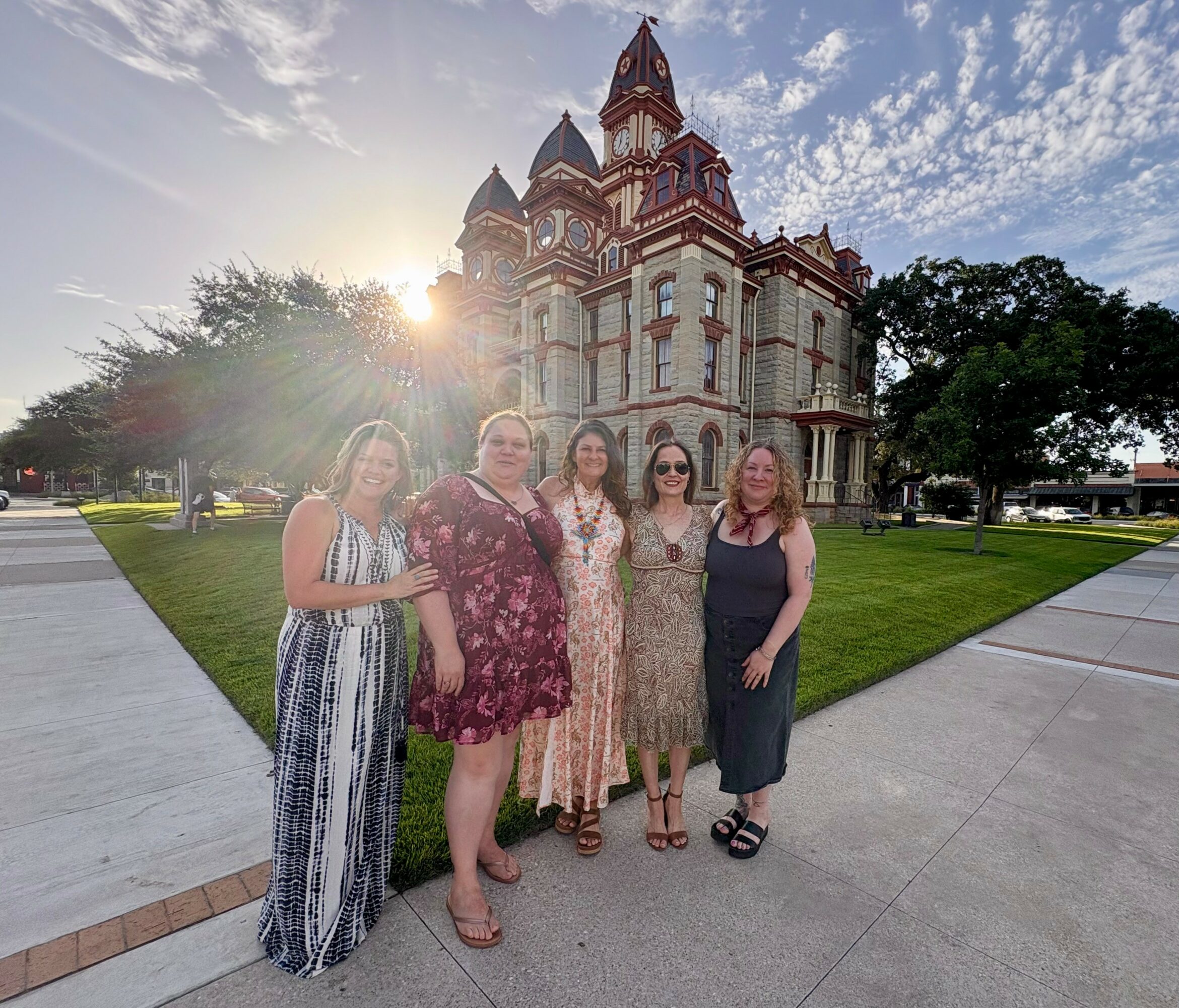 Five women stand in front of a large, ornate church with a bright sky and sun flare behind it.