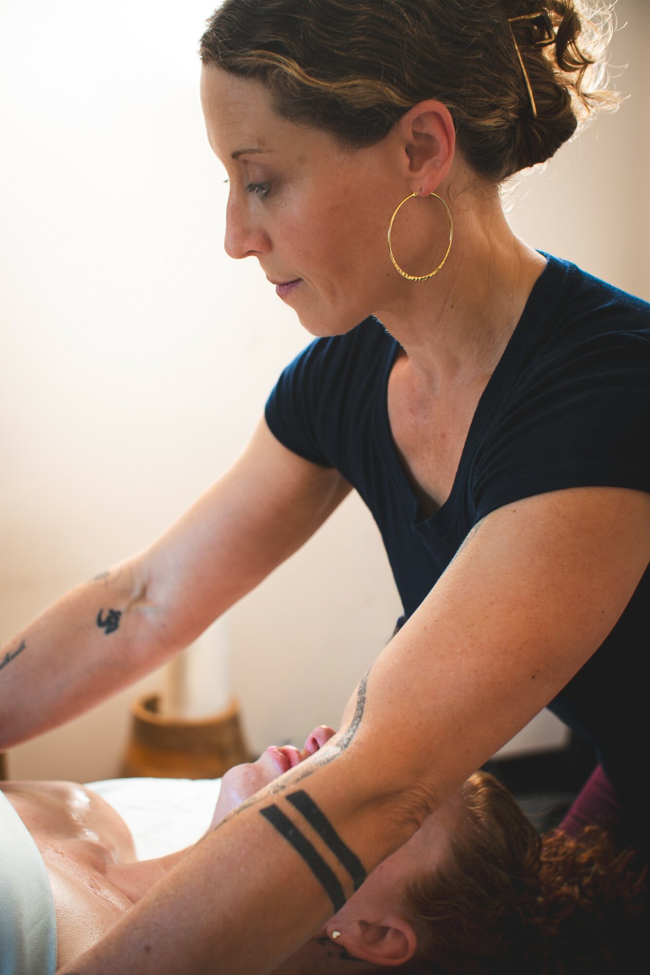 Woman with dark hair in a bun, wearing hoop earrings and a black shirt, tending to a person with tattoos on their arm.